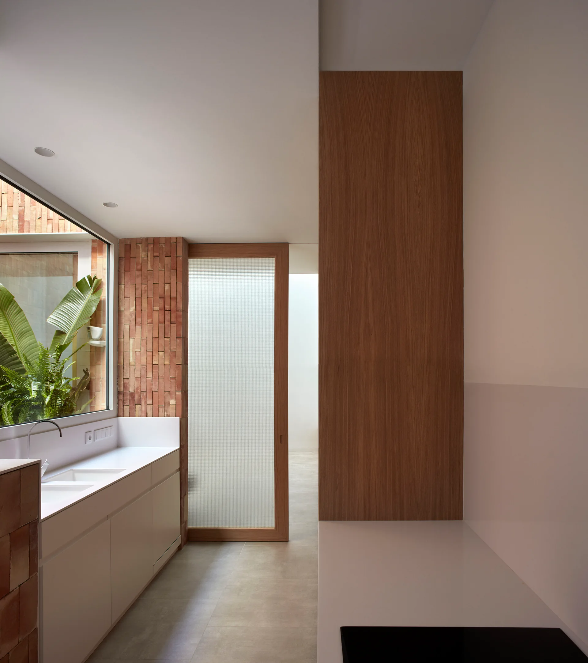 A minimalist kitchen features white cabinets, an undermount sink, a fluted glass sliding door, and a textured terracotta brick wall pillar.