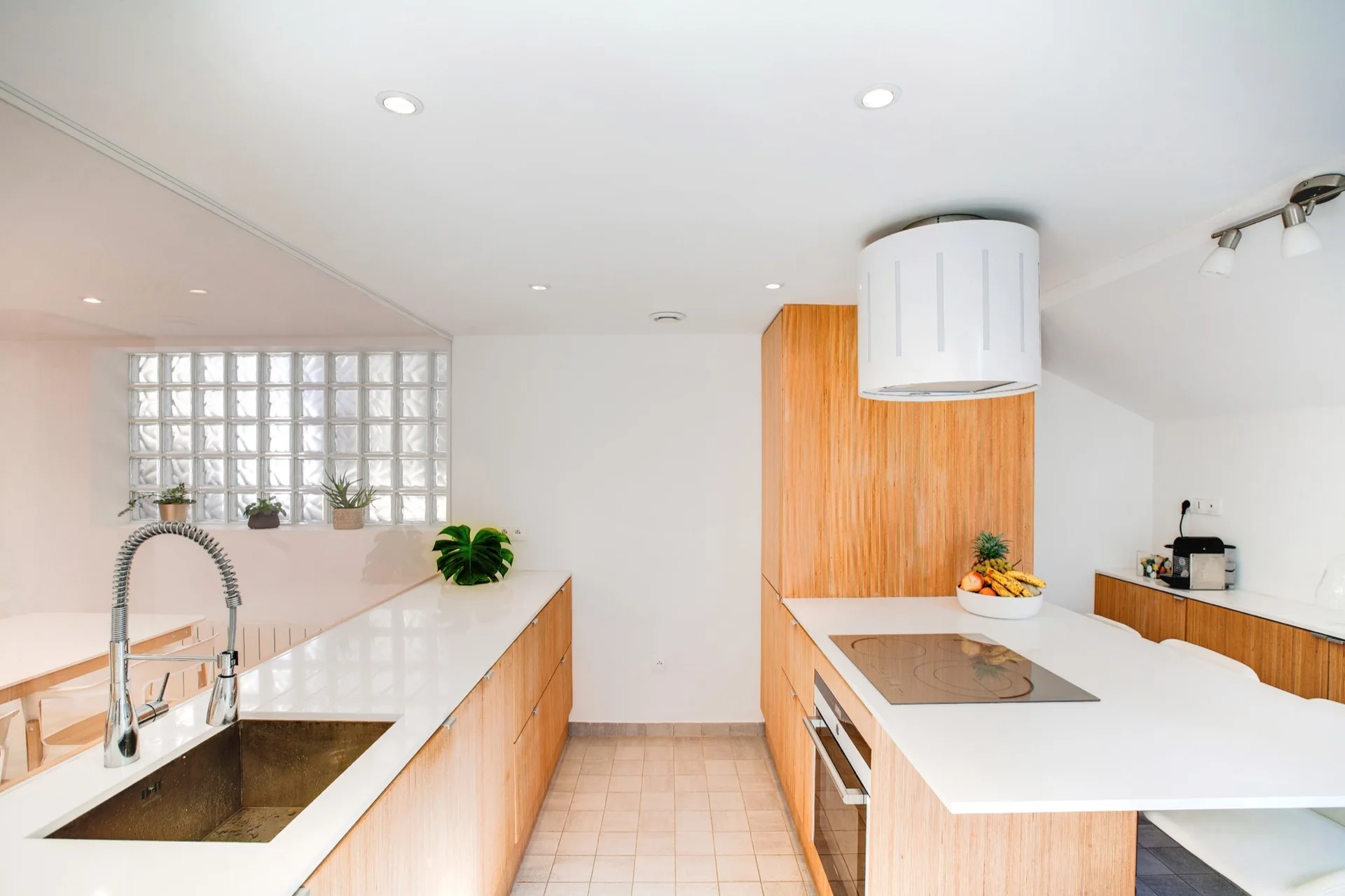 A modern kitchen featuring light wood cabinetry, white countertops, a central island with a cooktop, and a translucent glass block partition wall.