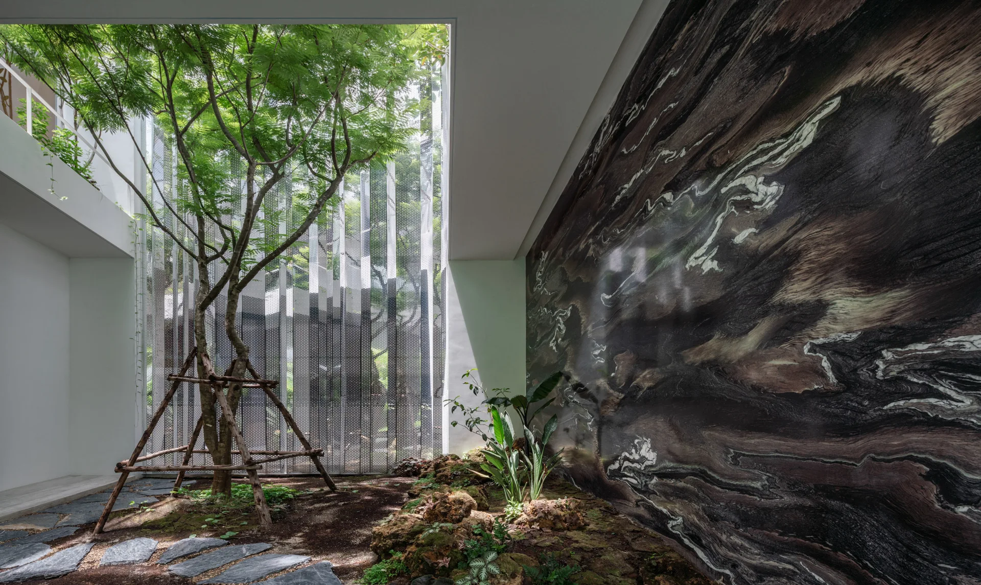 An indoor courtyard garden features a tall central tree, a dark polished marble-patterned feature wall, and a tall perforated metal screen.