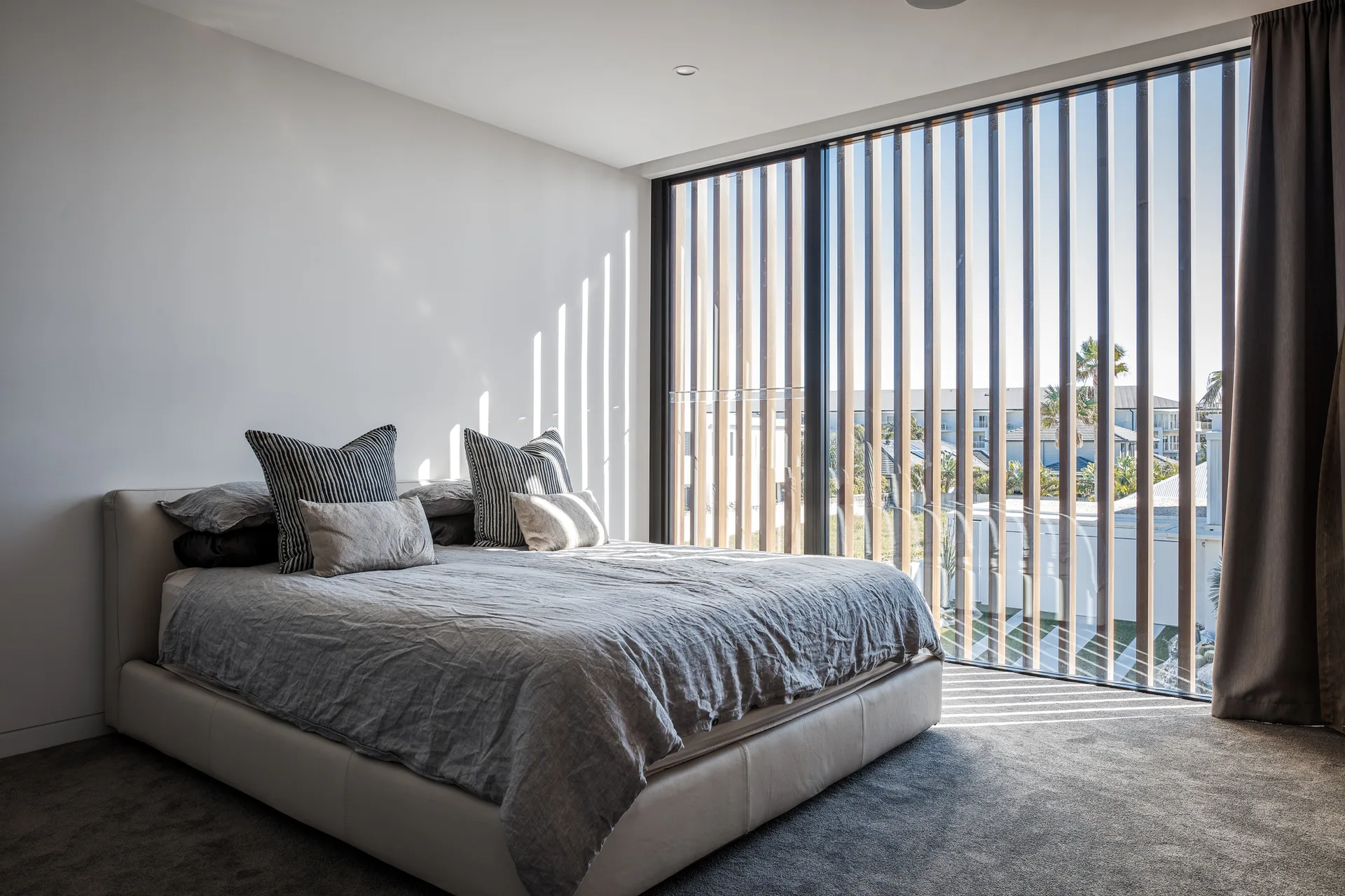 A minimalist bedroom features a low upholstered bed with grey linens and striped pillows next to a tall window with vertical wooden slats.