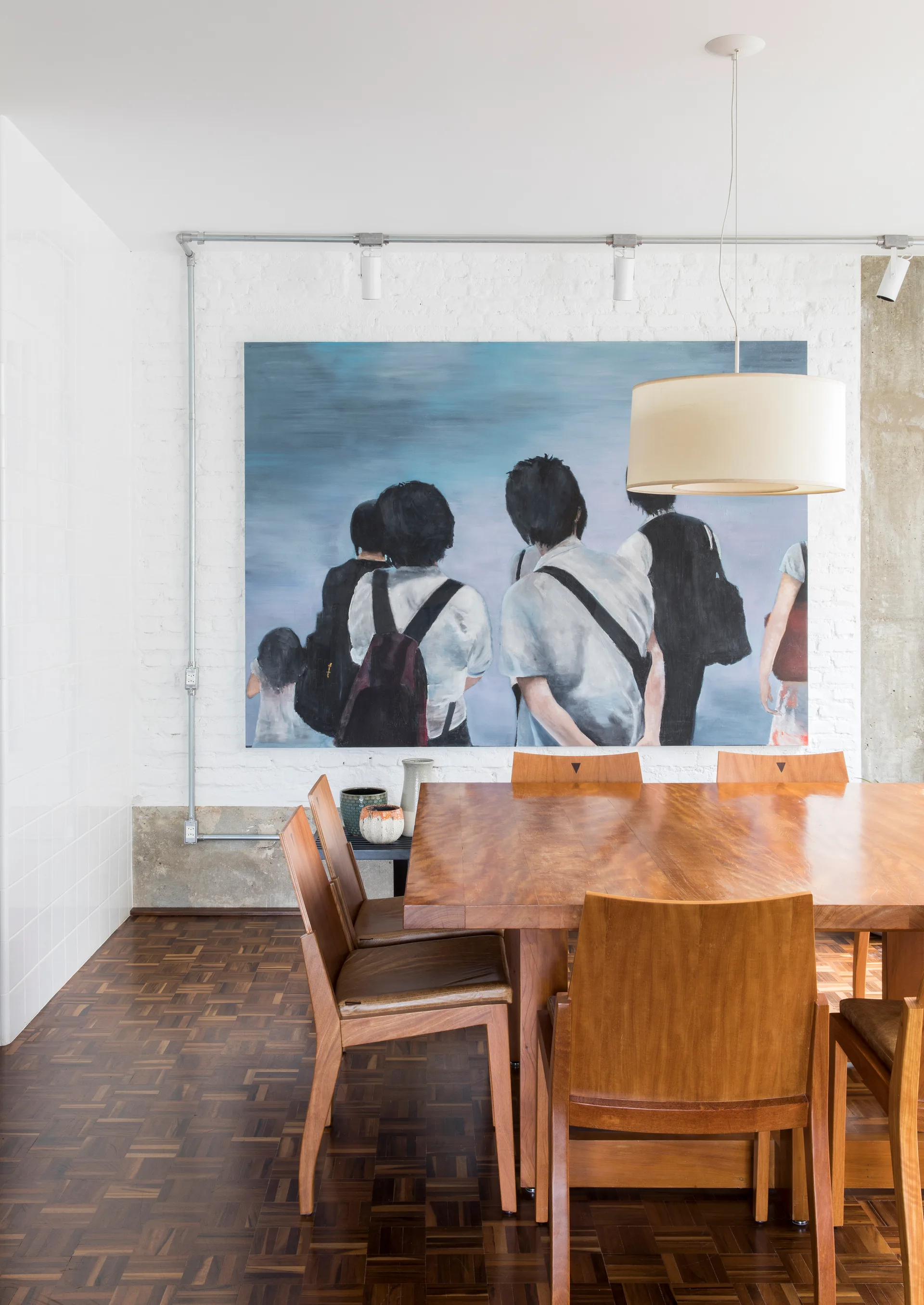 A wooden dining table and chairs set on parquet flooring in front of a white painted brick wall featuring a large blue figurative painting.