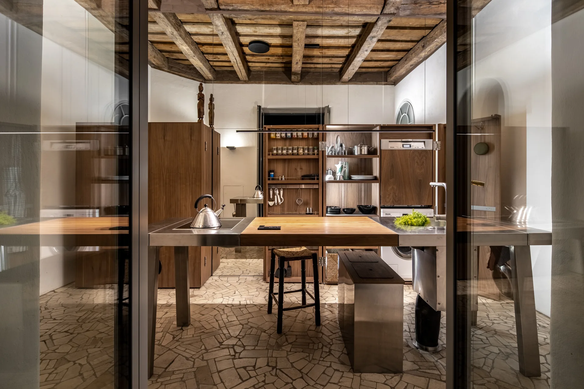 A modern kitchen features a central stainless steel island with a wood countertop under an exposed timber ceiling and irregular stone flooring.