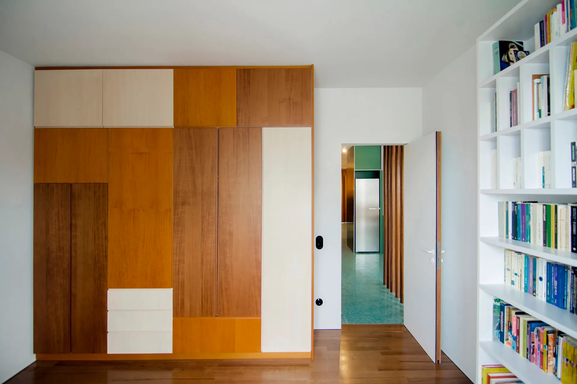 A minimalist room features a multi-toned wood wardrobe wall and a white recessed bookshelf next to an open doorway showing a green tiled hallway.