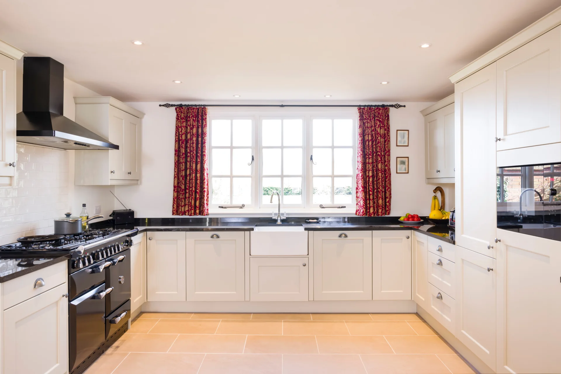A U-shaped cream kitchen features a black range stove, black countertops, a white farmhouse sink beneath a large window, and patterned red curtains.