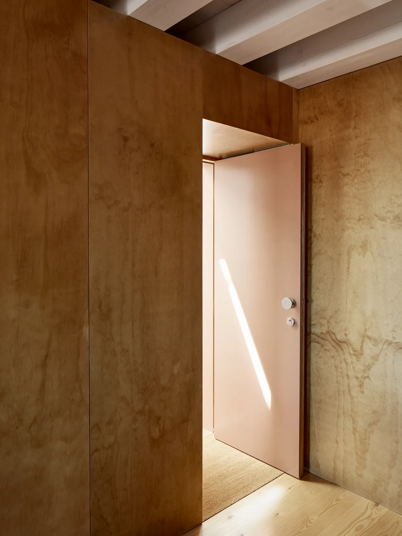 A minimalist hallway features warm plywood wall paneling, light wood flooring, and a partially open peach-colored door under white ceiling beams.