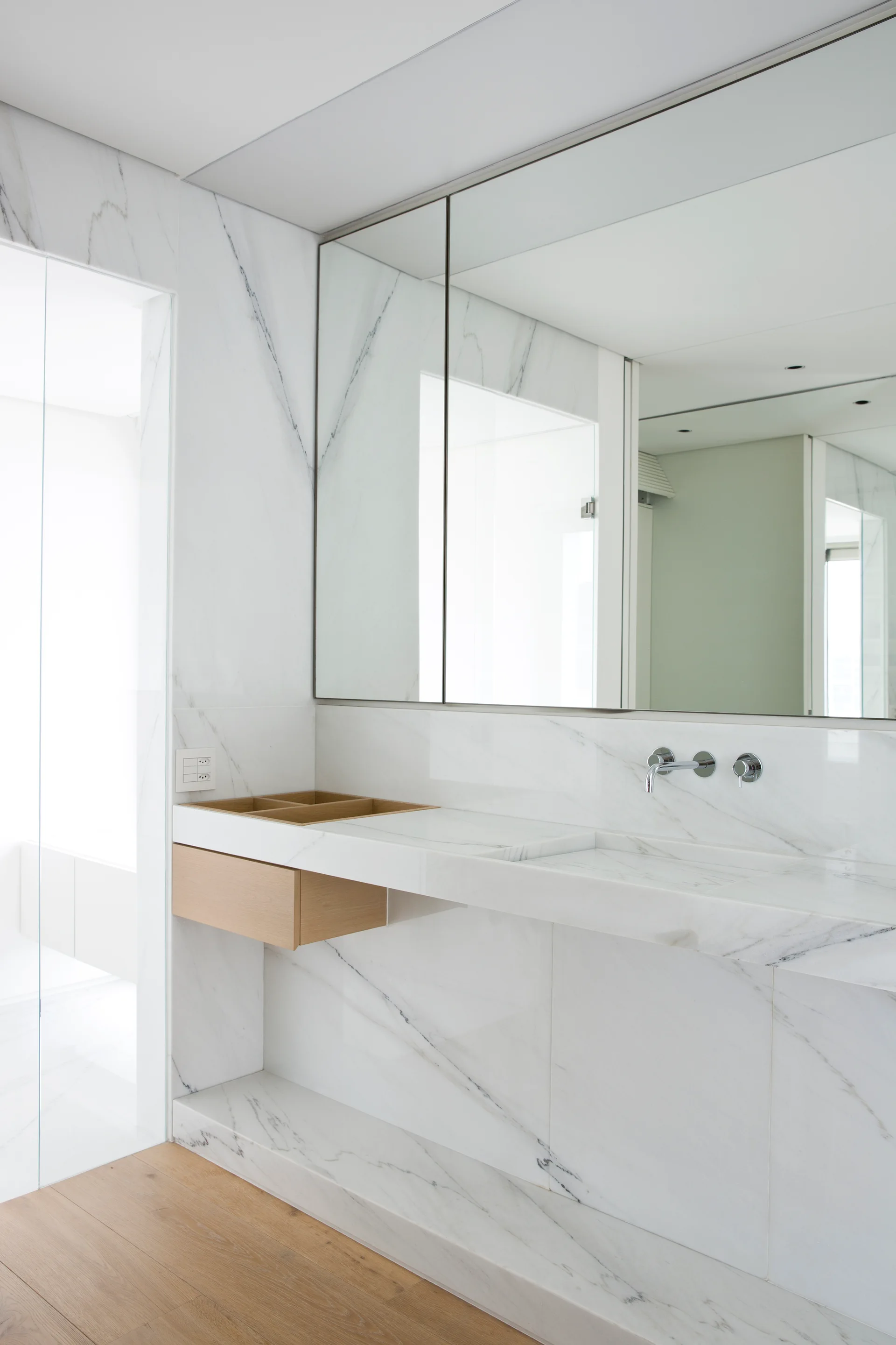 A modern bathroom features a white marble vanity with an integrated sink, a wall-mounted faucet, a large frameless mirror, and a floating wood drawer.