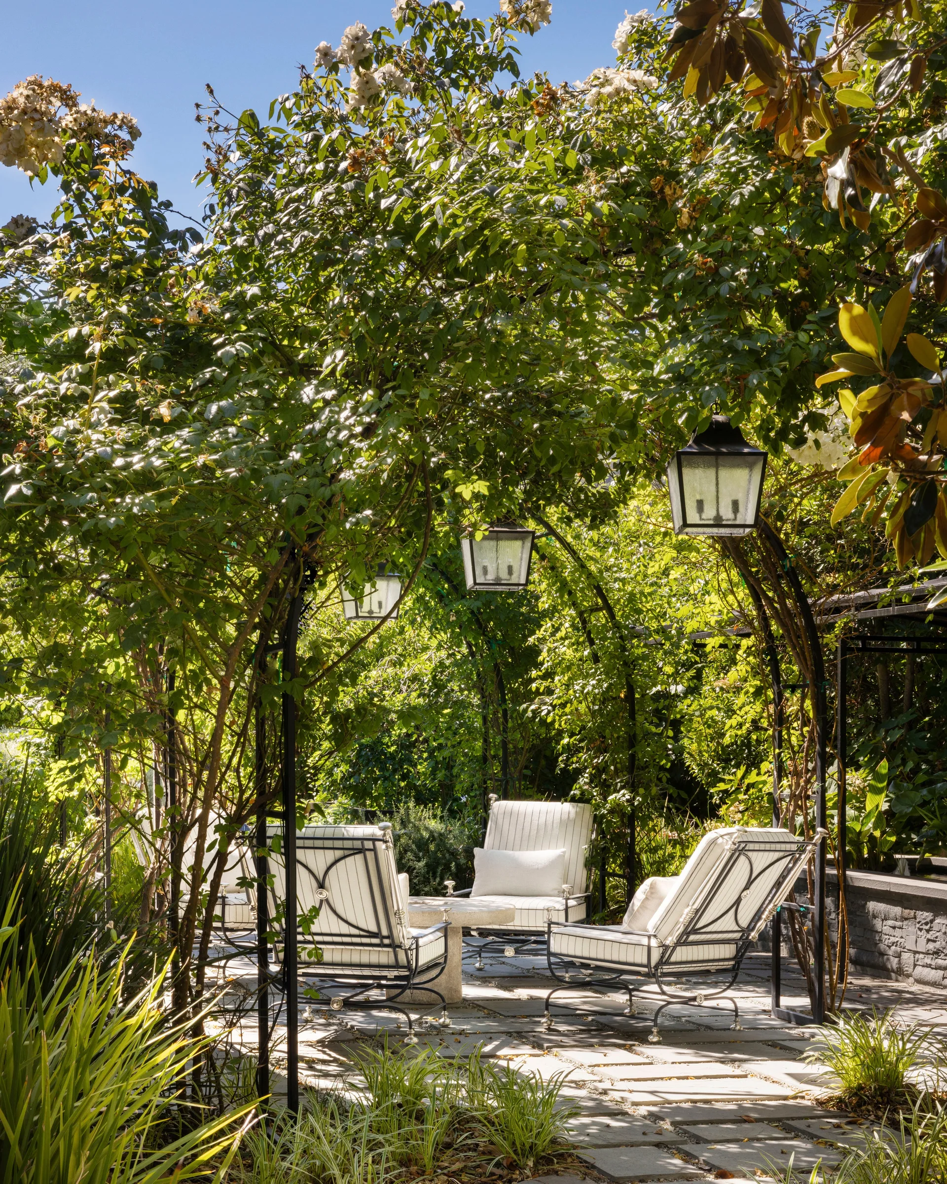 Black wrought iron lounge chairs with striped cushions sit on a stone paver patio beneath a lush green arched trellis hung with black lanterns.
