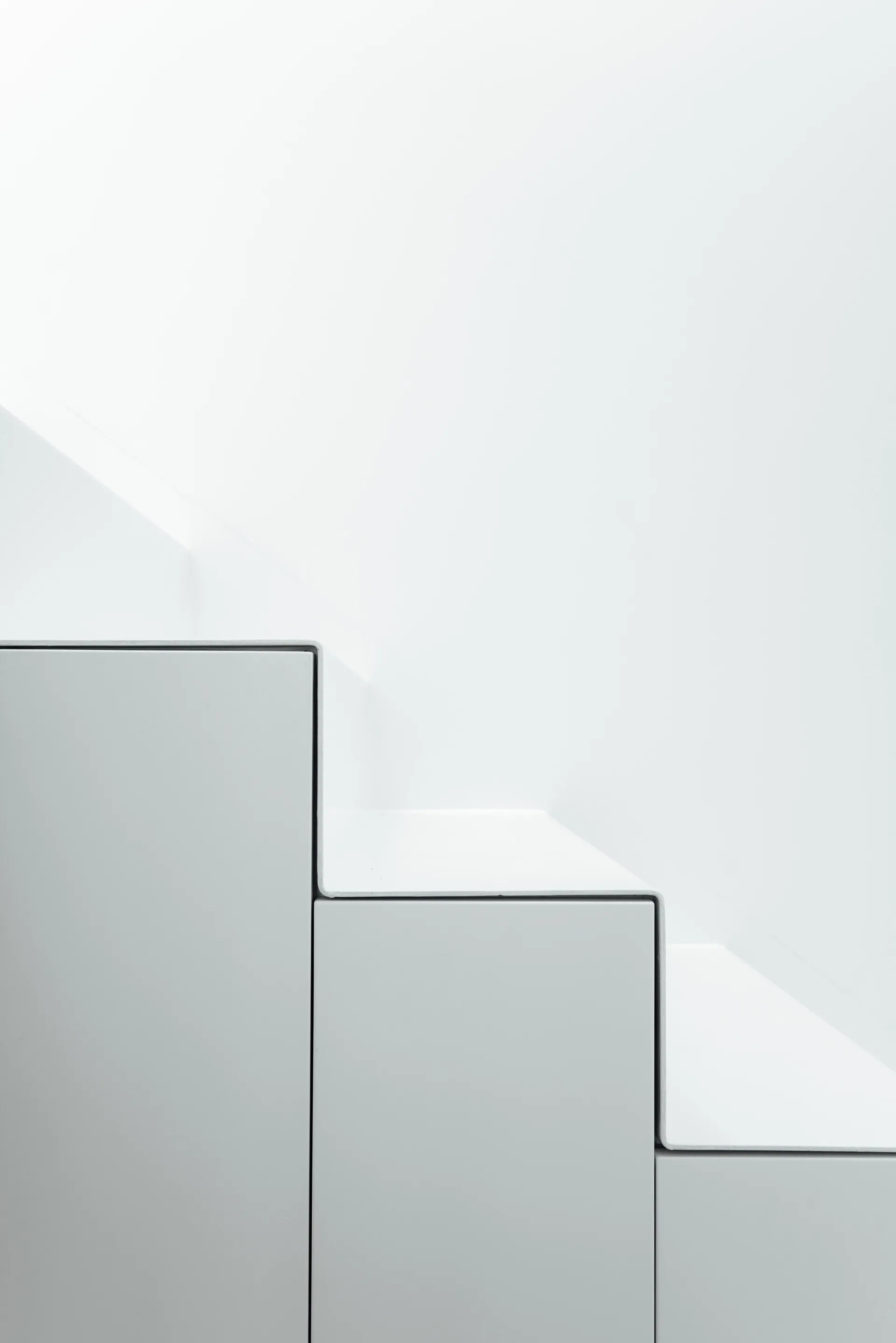 A close-up view of a minimalist white metal staircase showing sharp geometric treads and risers against a plain white wall backdrop.