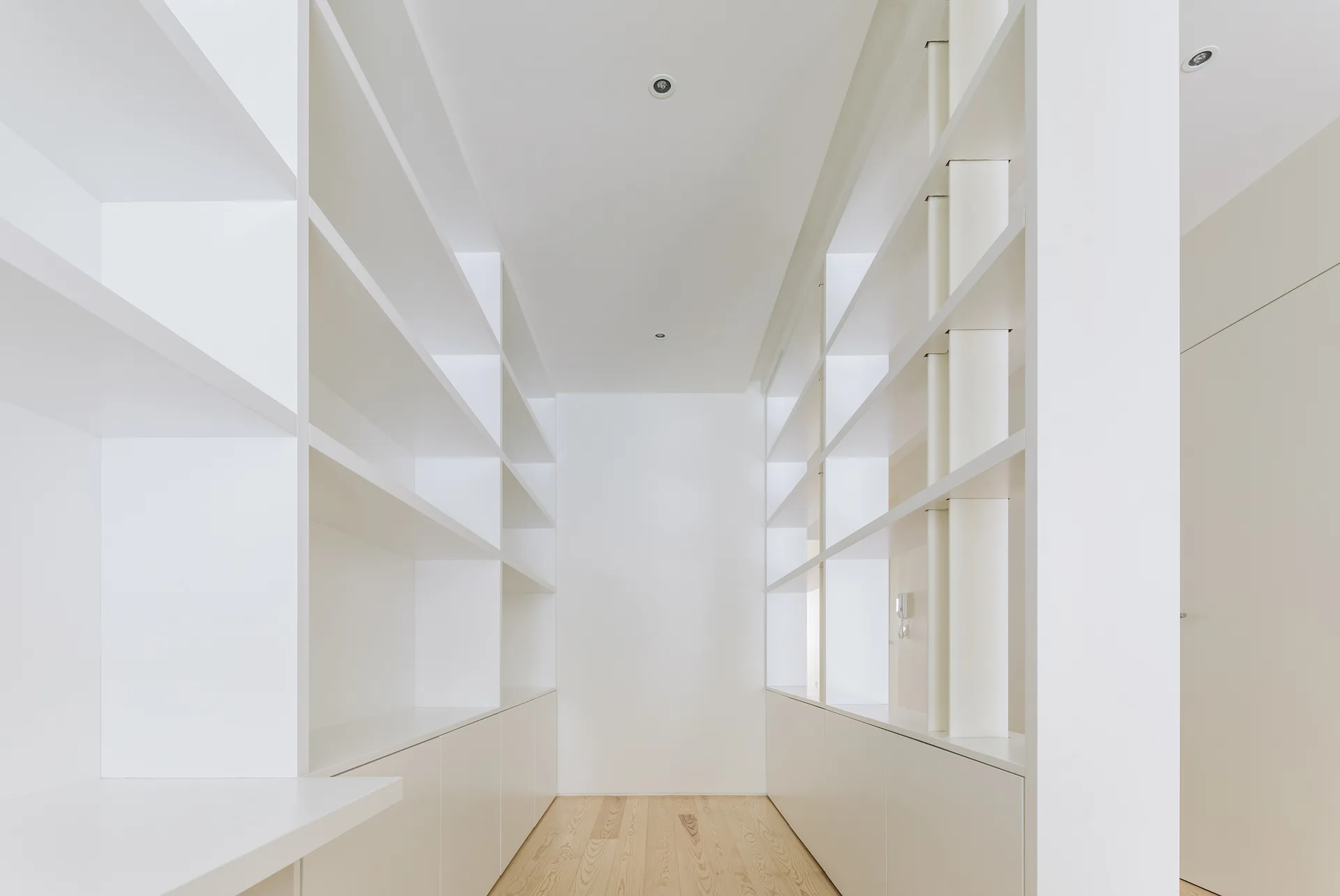 A minimalist walk-in closet featuring floor-to-ceiling white built-in shelving, base cabinets, and light wood flooring under recessed ceiling lights.
