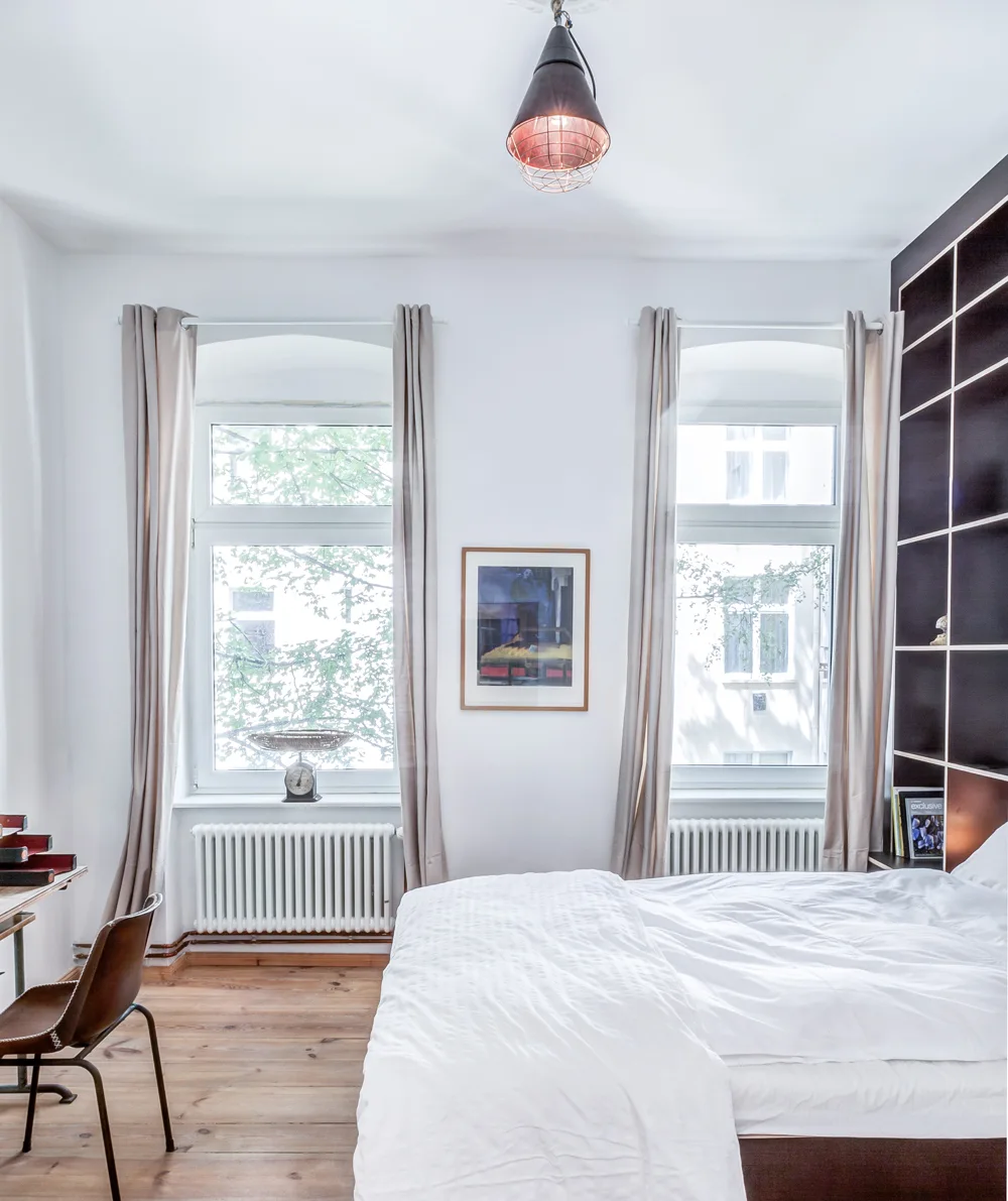 A minimalist white bedroom with symmetrical windows, radiators, and a tall dark grid bookshelf next to a bed with white bedding.