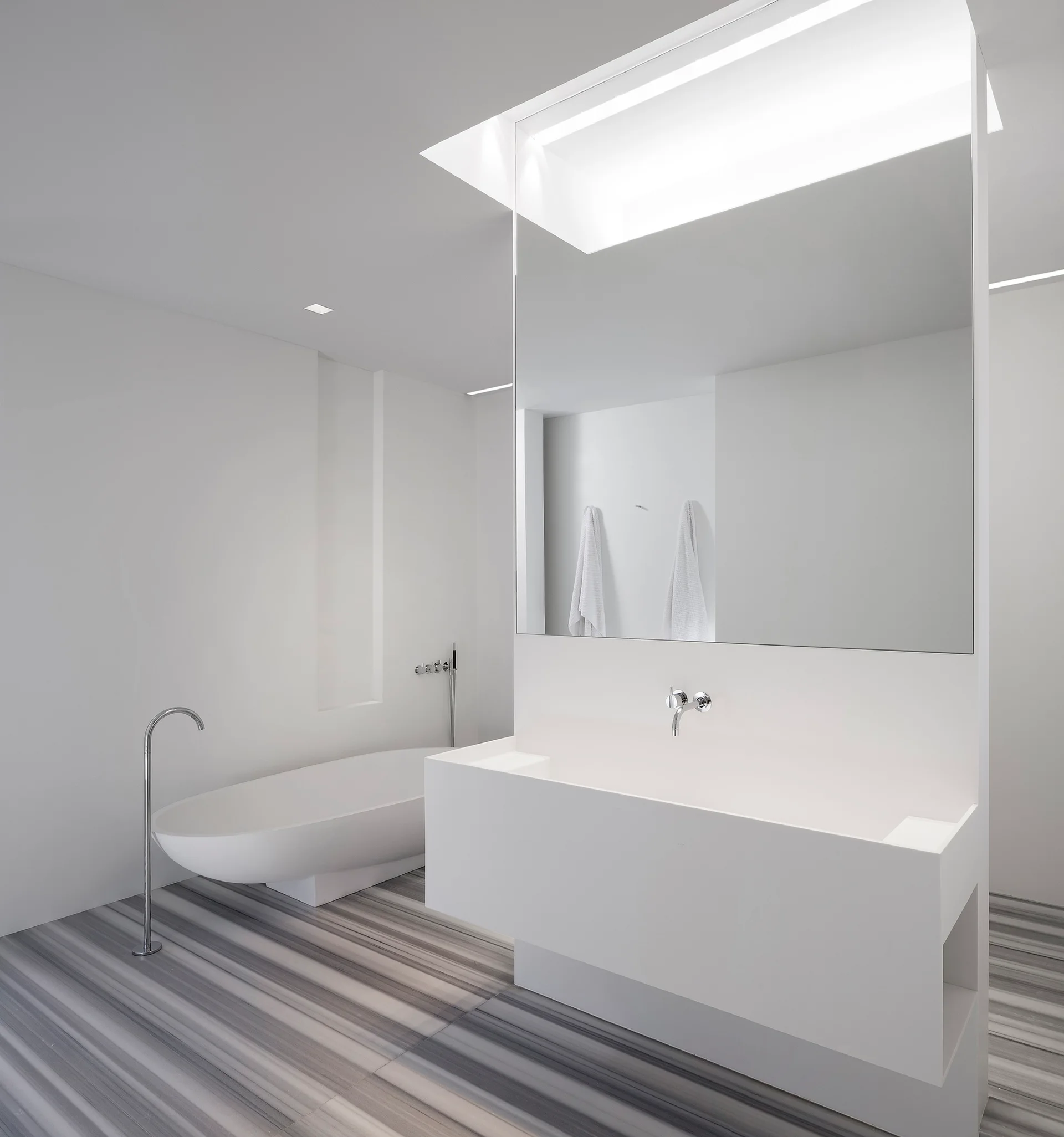 A minimalist white bathroom featuring a large rectangular block vanity, a freestanding oval tub, and grey linear-striped marble flooring.