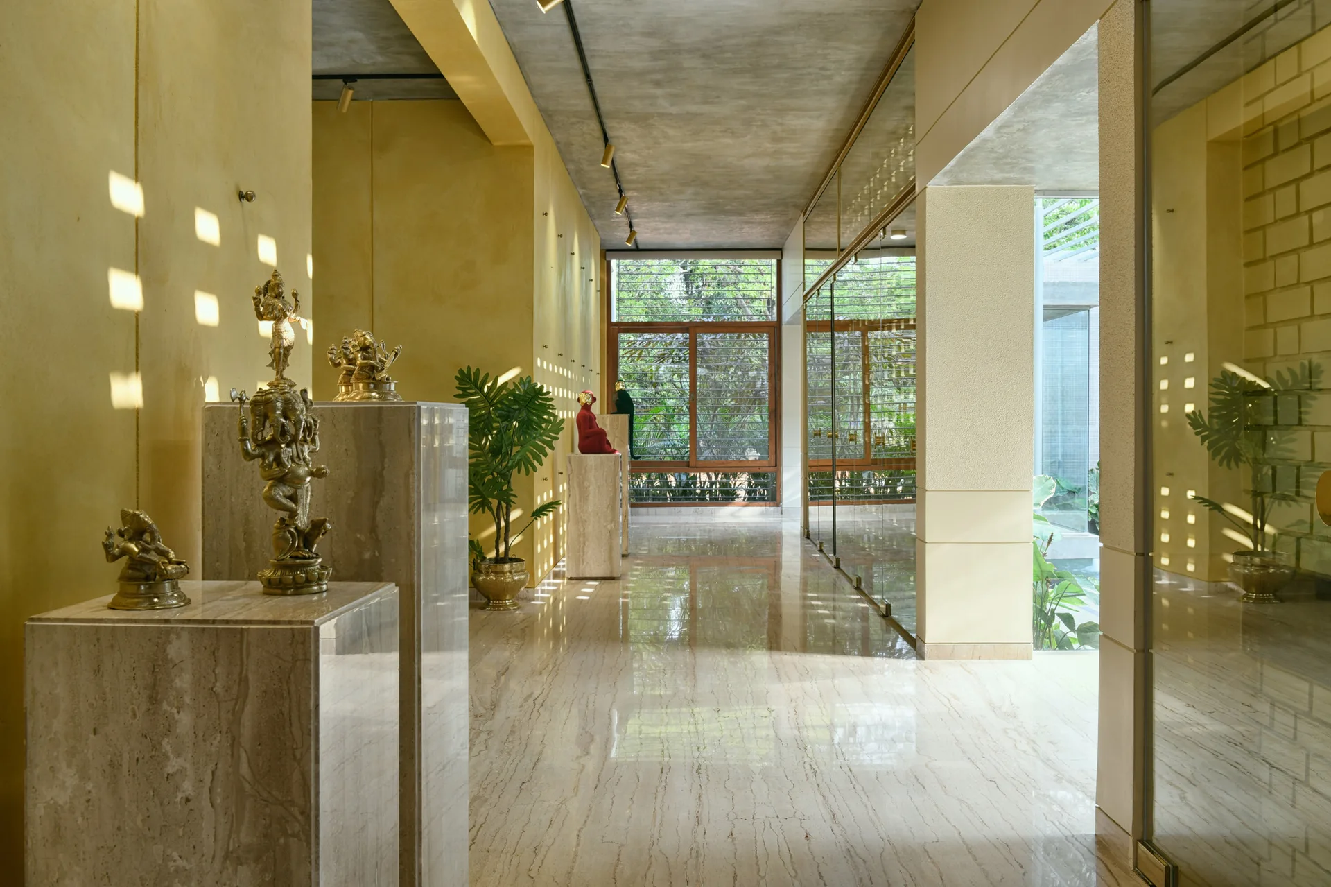 A long hallway features warm yellow walls, a polished beige marble floor, and stone display pedestals holding golden sculptures below a concrete ceiling.