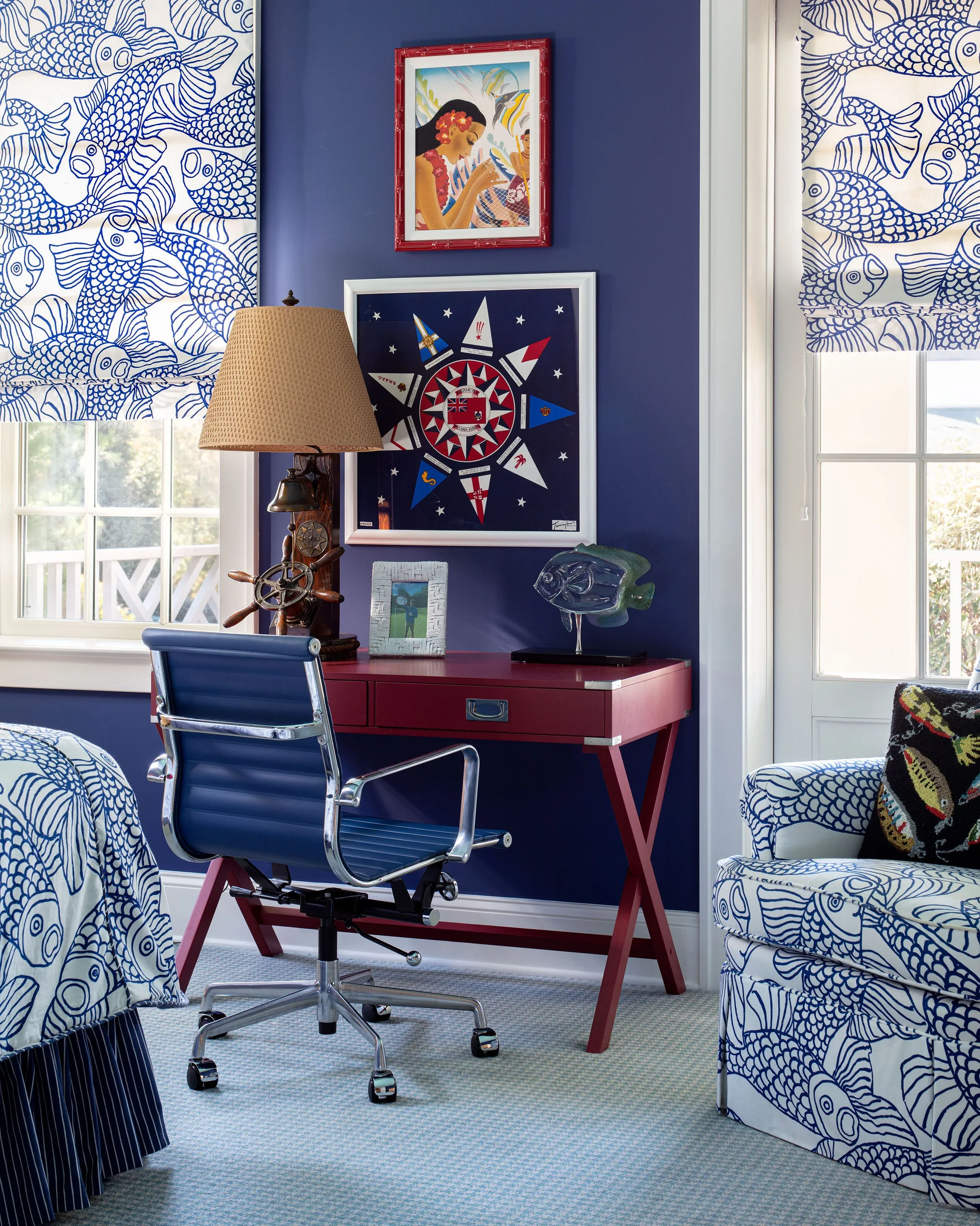 A red writing desk and blue office chair stand against a dark navy wall next to windows with blue and white fish pattern shades.