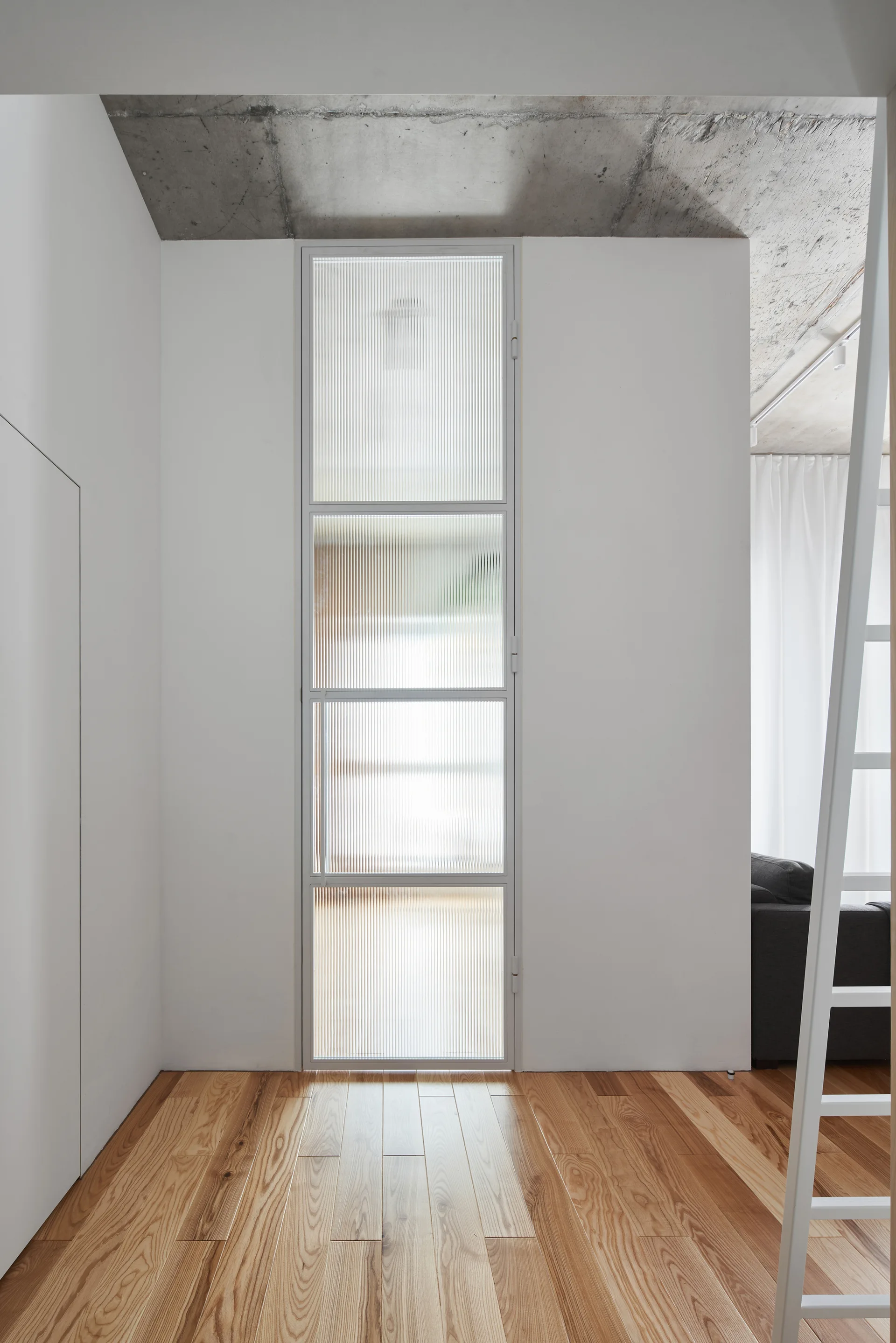 A minimalist hallway features a tall fluted glass door panel, light wood plank flooring, and an exposed concrete ceiling.