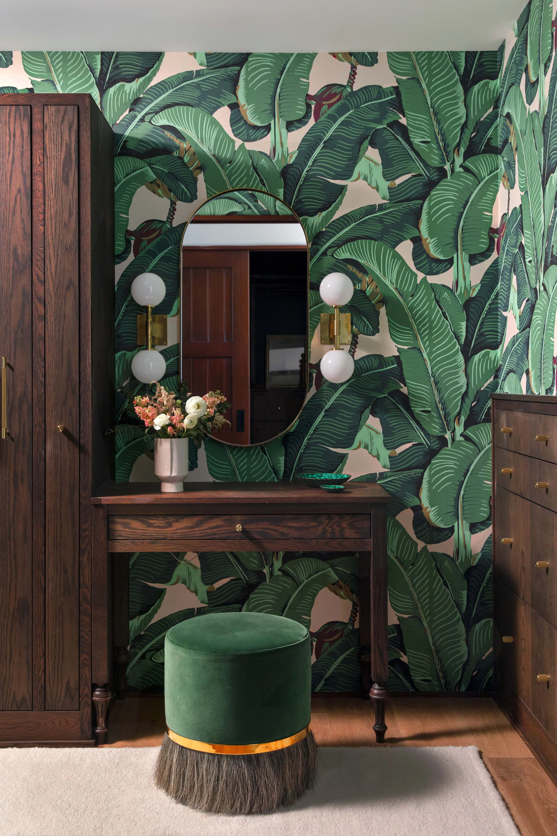 A dark wood vanity table with a green velvet stool sits against a bold green banana leaf wallpapered wall, flanked by matching wood cabinets.