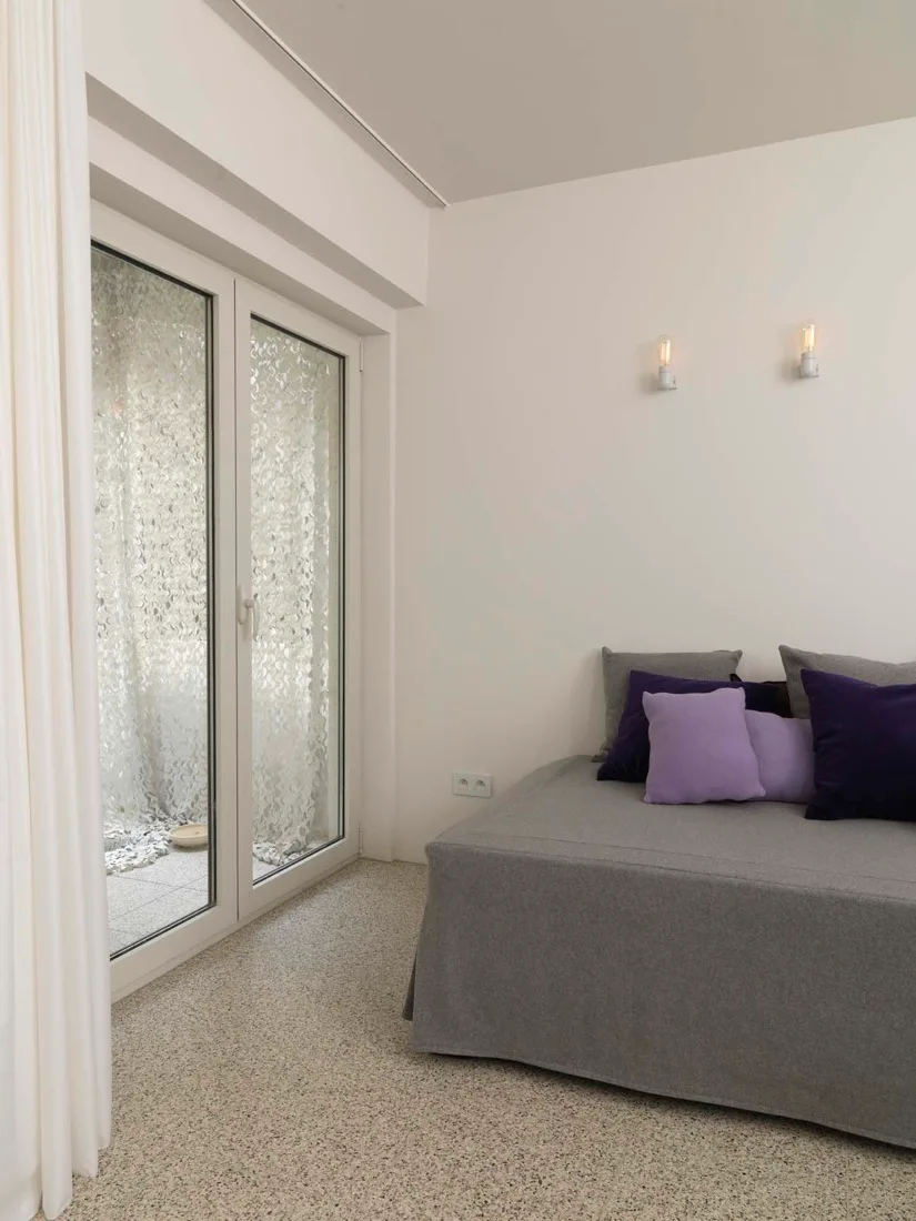 A minimalist bedroom featuring a grey slipcovered daybed with purple pillows against white walls with two bare-bulb sconces.