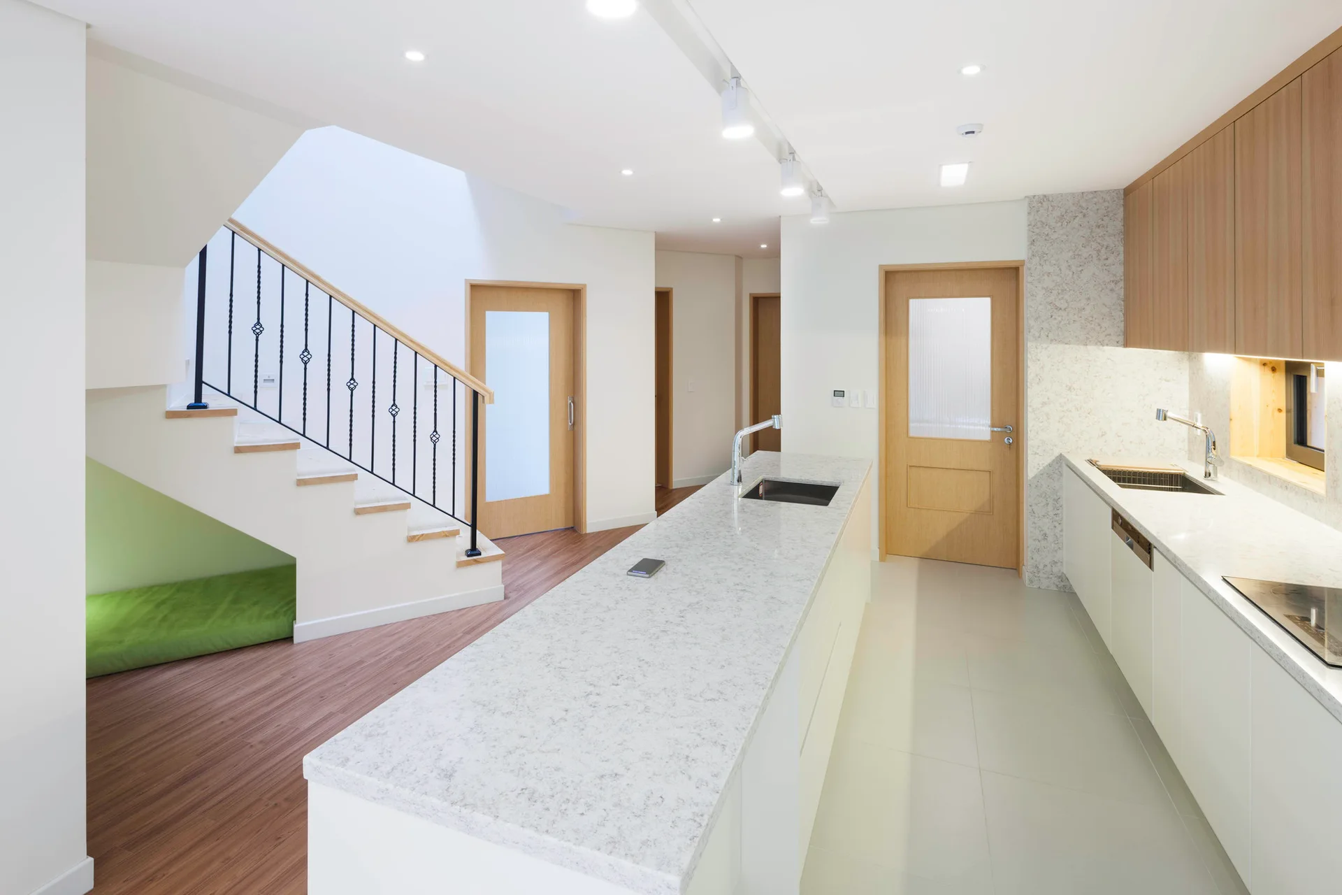 A bright minimalist kitchen featuring a long speckled white island, wood cabinets, and an adjacent white staircase with a green cushion nestled underneath.