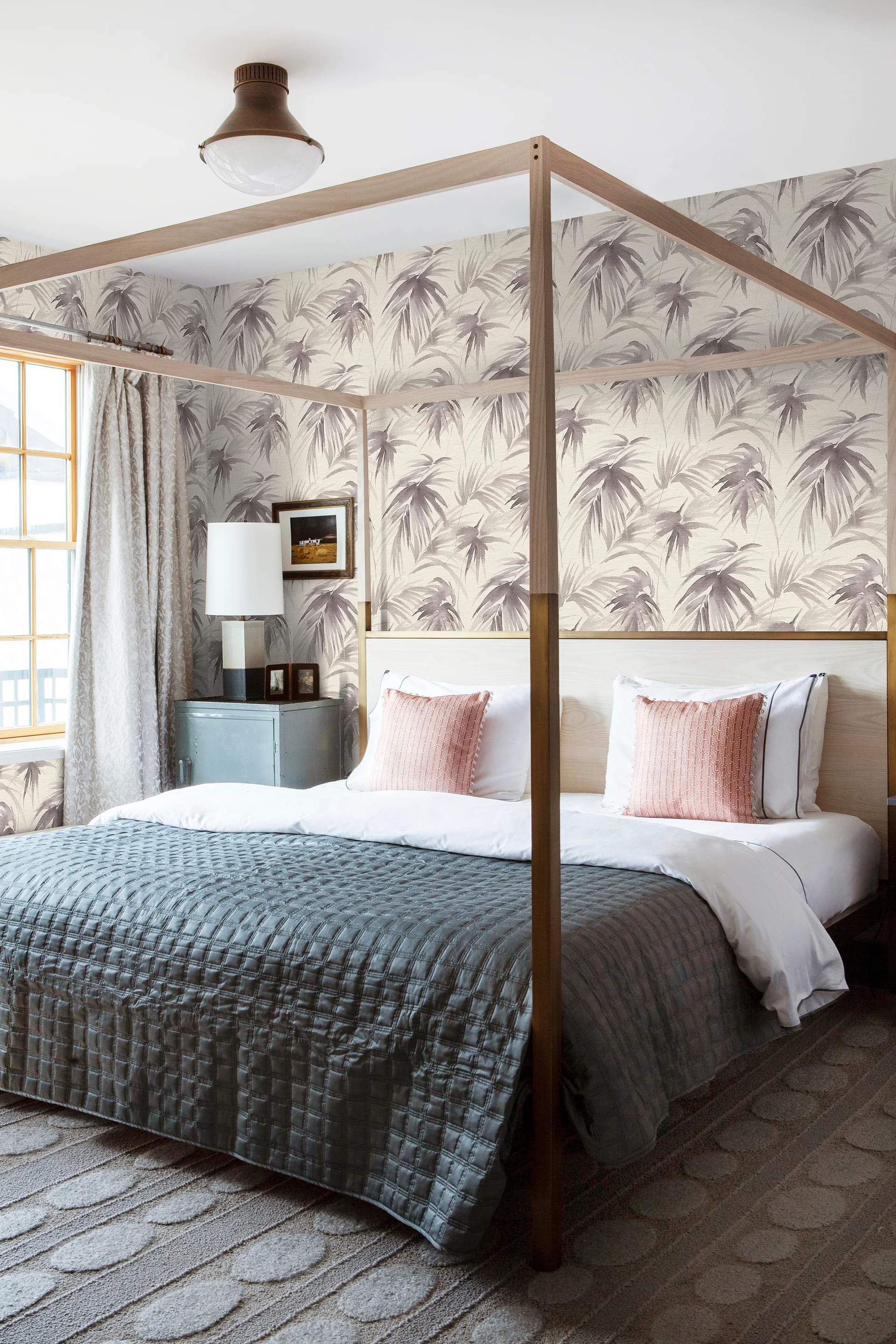 A bedroom features a wood-framed canopy bed, grey quilted bedding, pink accent pillows, and palm-leaf patterned wallpaper on the walls.
