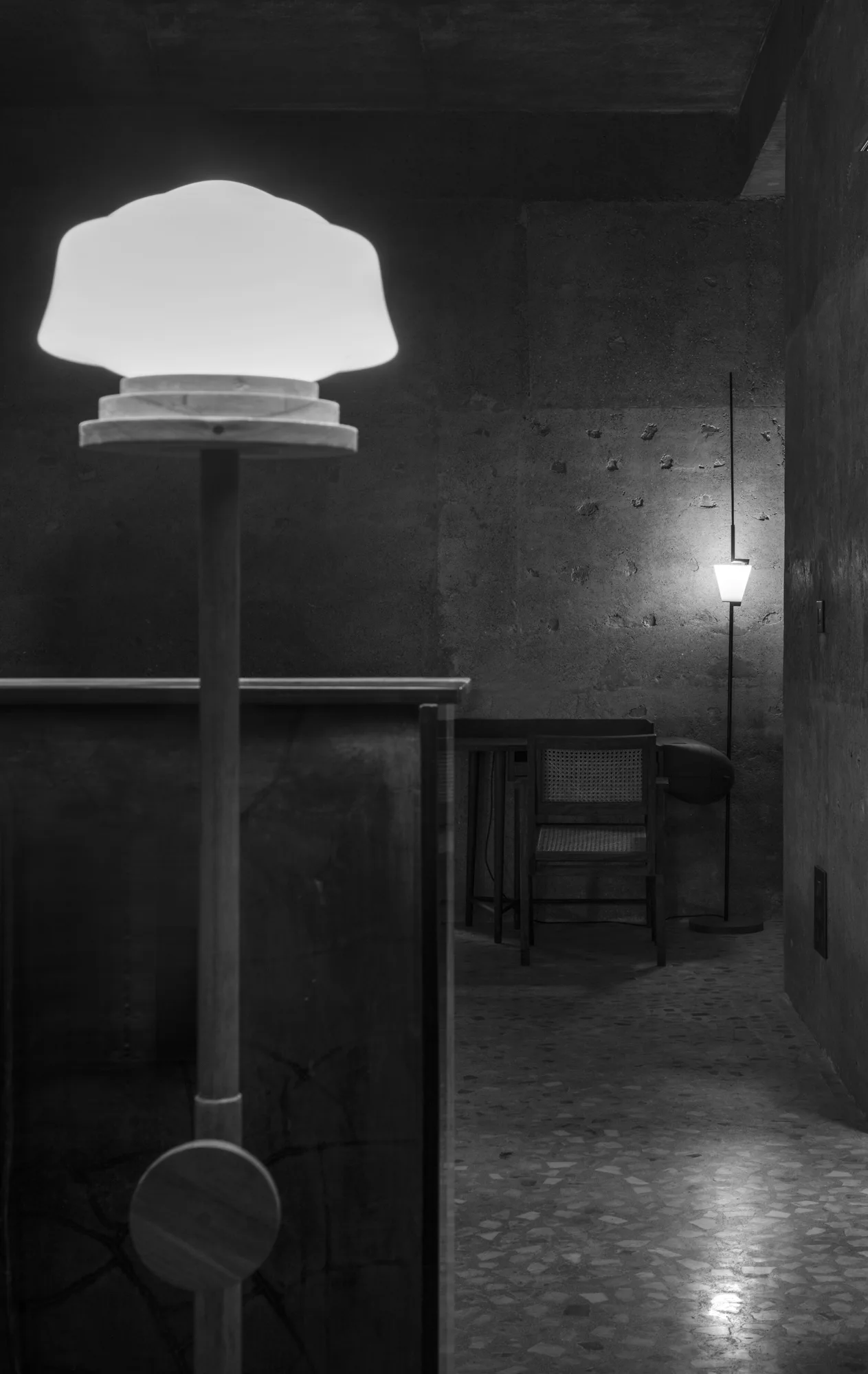 Illuminated floor lamps cast a soft glow over textured concrete walls, a cane back chair, and speckled terrazzo flooring in a dimly lit room.