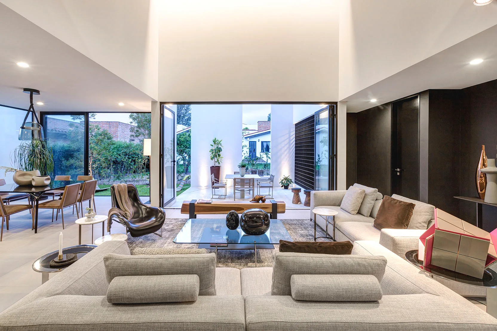A spacious modern living room with a grey modular sofa, a glass coffee table, and folding glass doors that open to a courtyard.