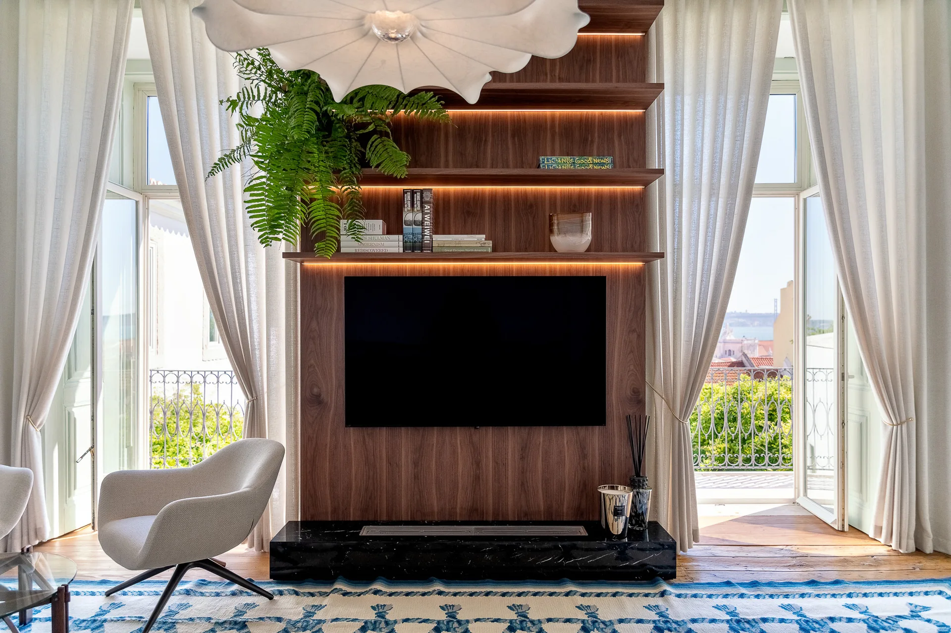 A dark wood built-in media wall features a TV, backlit shelves with a fern, and a black stone hearth, flanked by floor-to-ceiling cream curtains.