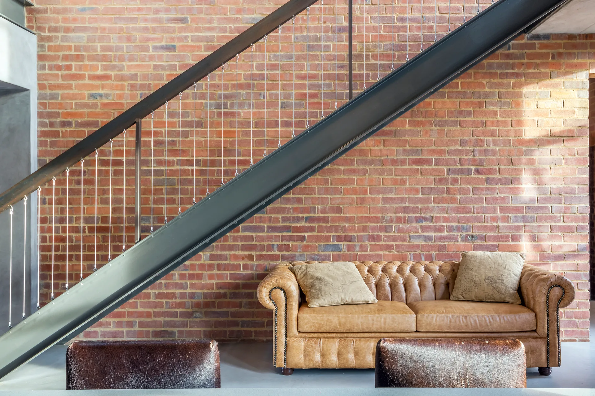 A tan leather Chesterfield sofa sits beneath an industrial steel staircase with cable railings against an exposed red brick wall.