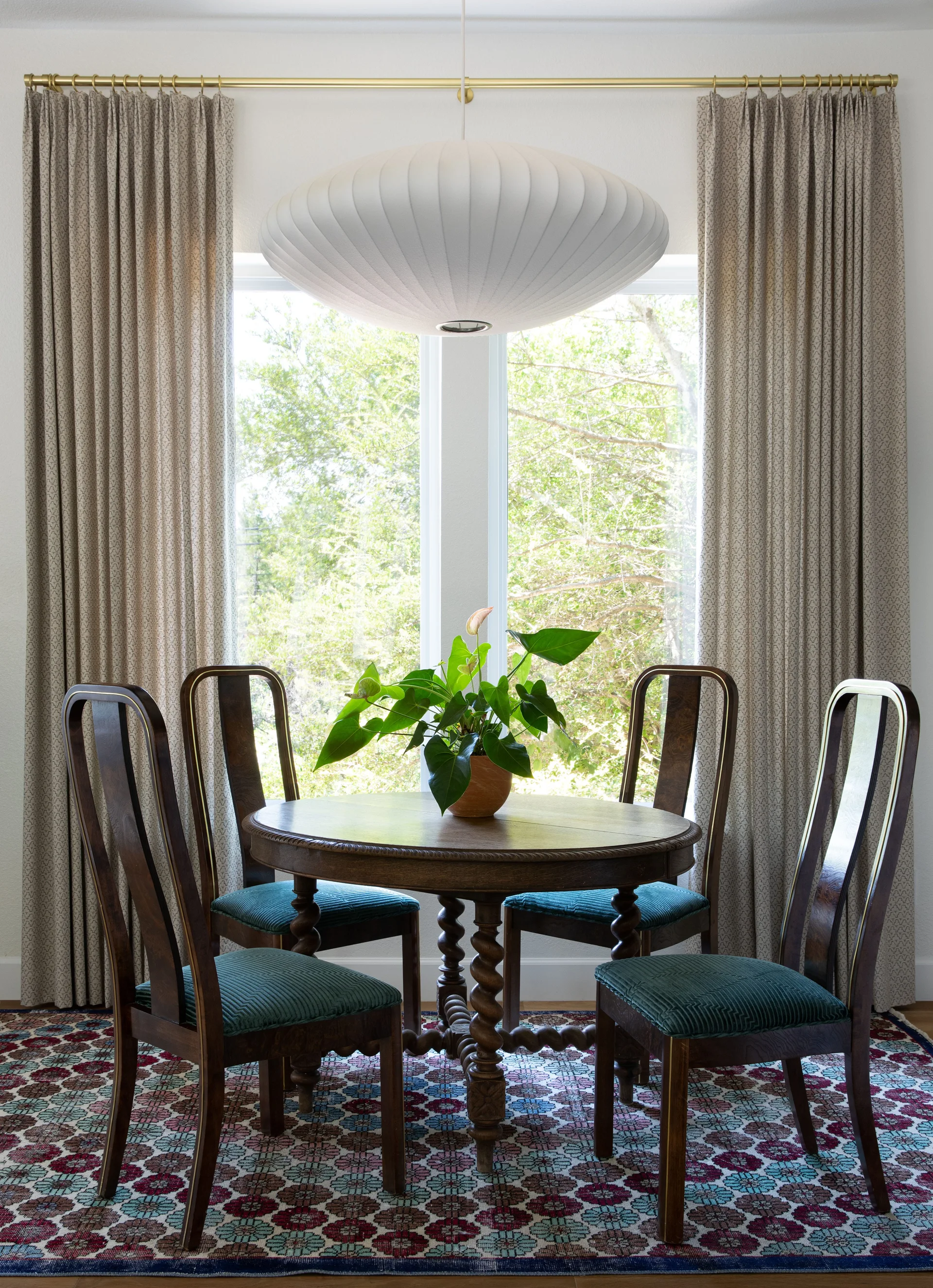 Four dark wood chairs with green upholstered seats surround a round wooden pedestal table under a white saucer pendant light in front of a window.