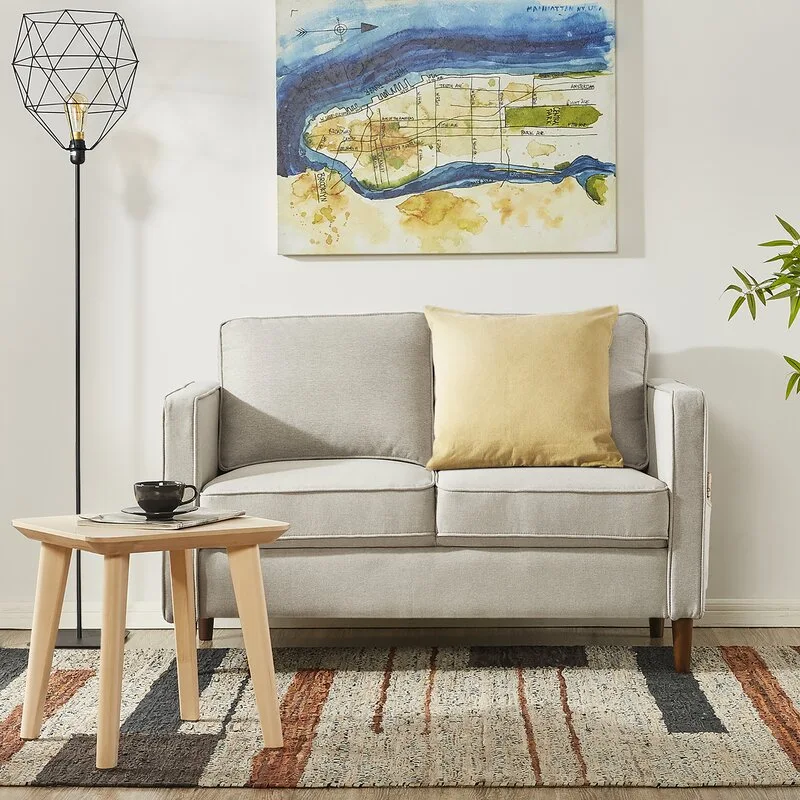 A light gray two-seater sofa with a yellow pillow sits on a woven patterned rug next to a light wood side table and a black cage floor lamp.