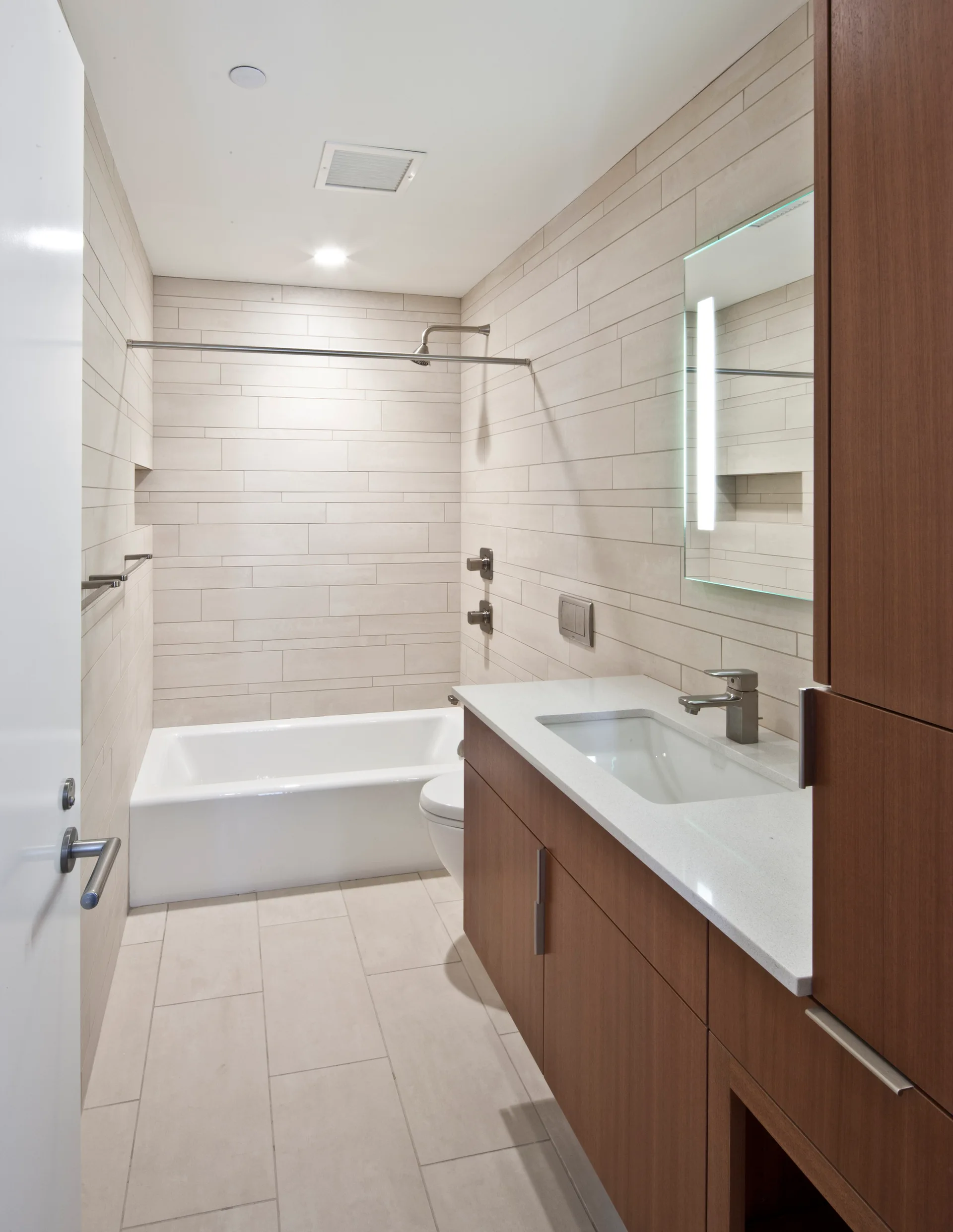 A modern bathroom featuring horizontal beige wall tiles, a wood-toned vanity with a white countertop, an illuminated mirror, and a white bathtub.