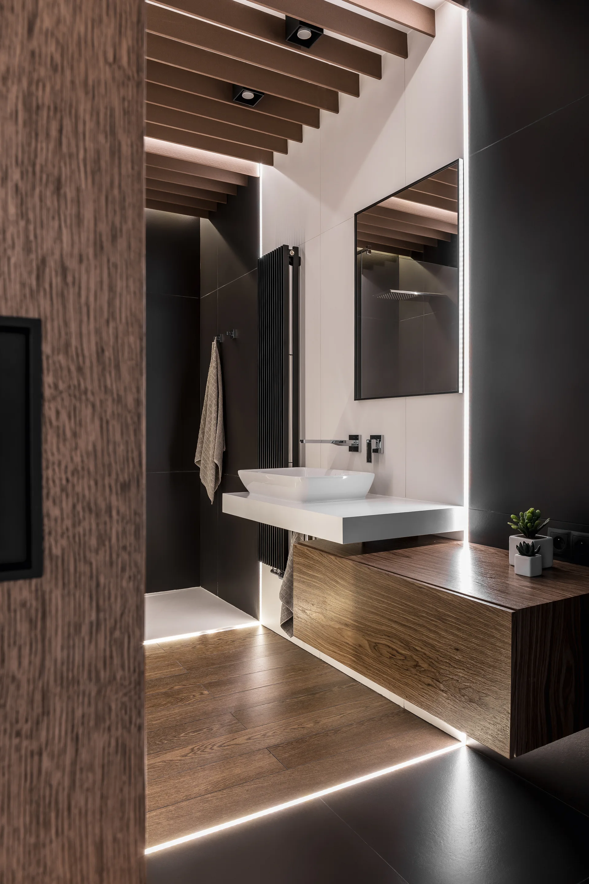 A modern bathroom features a floating wooden vanity, white vessel sink, rectangular mirror, wood-slatted ceiling, and integrated LED light strips.