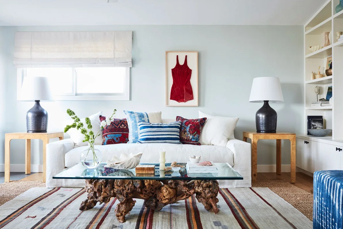 A light blue living room features a white slipcovered sofa, a root wood coffee table with a glass top, and symmetrical dark table lamps.