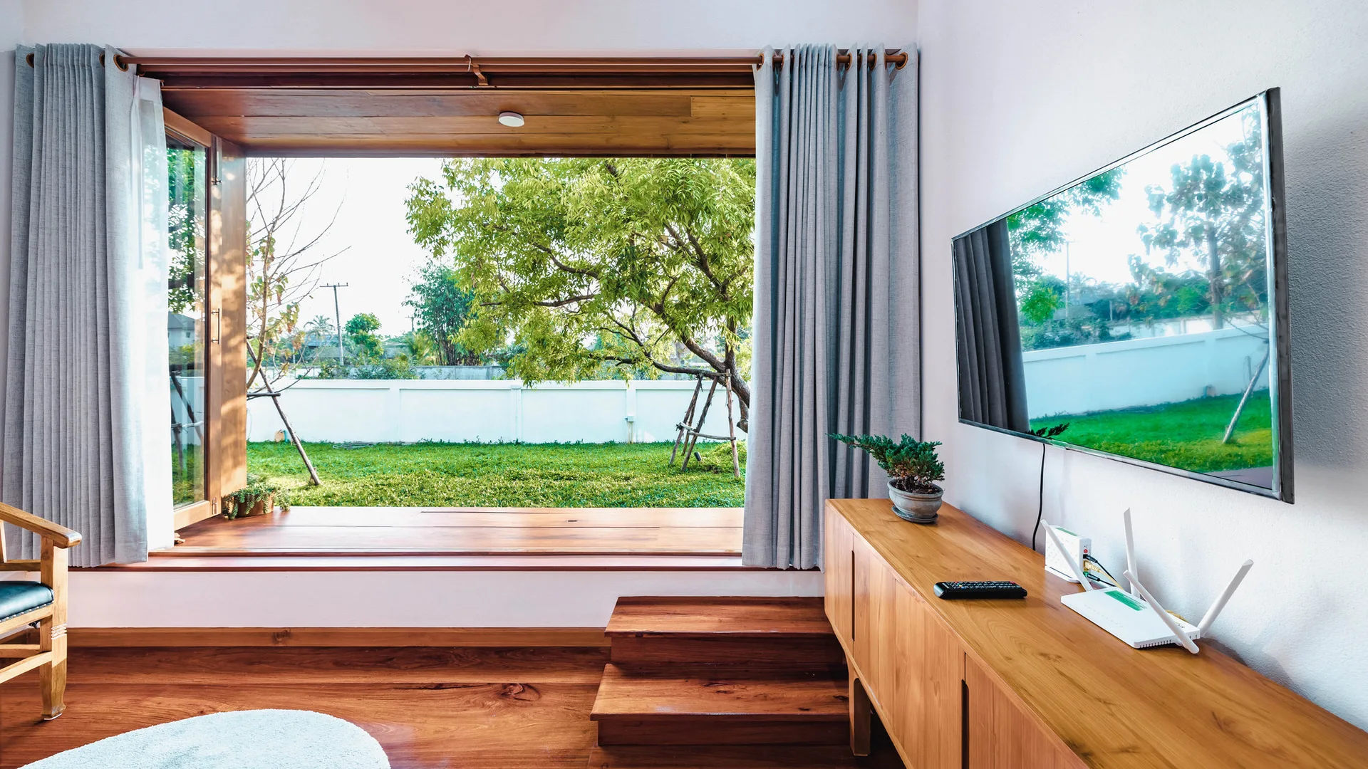 A minimalist living room features a large wood-framed window looking onto a green lawn, next to a wooden sideboard and wall-mounted TV.