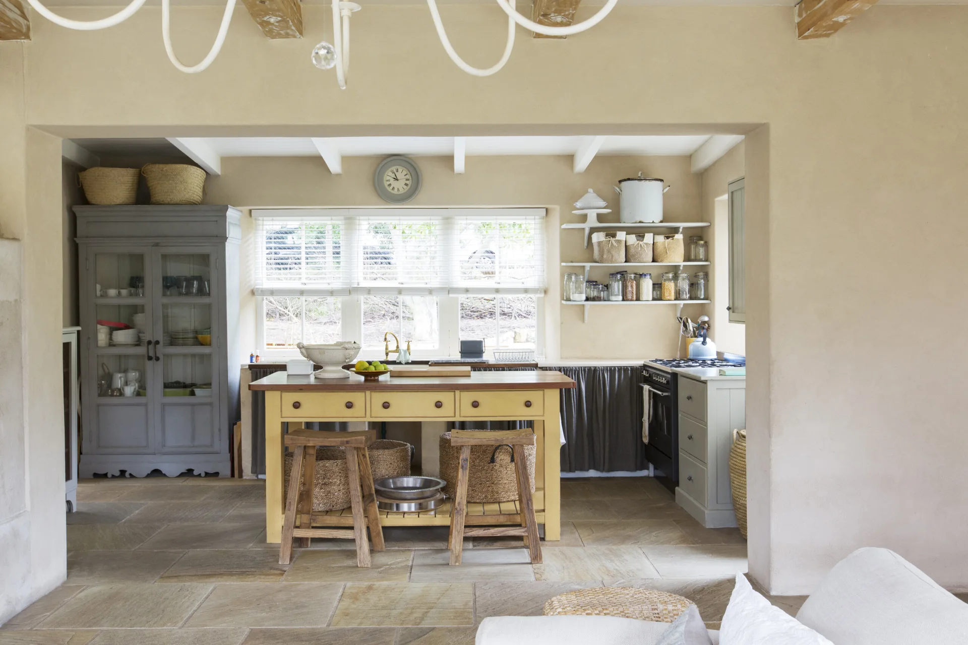 A rustic kitchen features a yellow central island with wooden stools, a tall grey storage cabinet, and beige natural stone tile flooring.