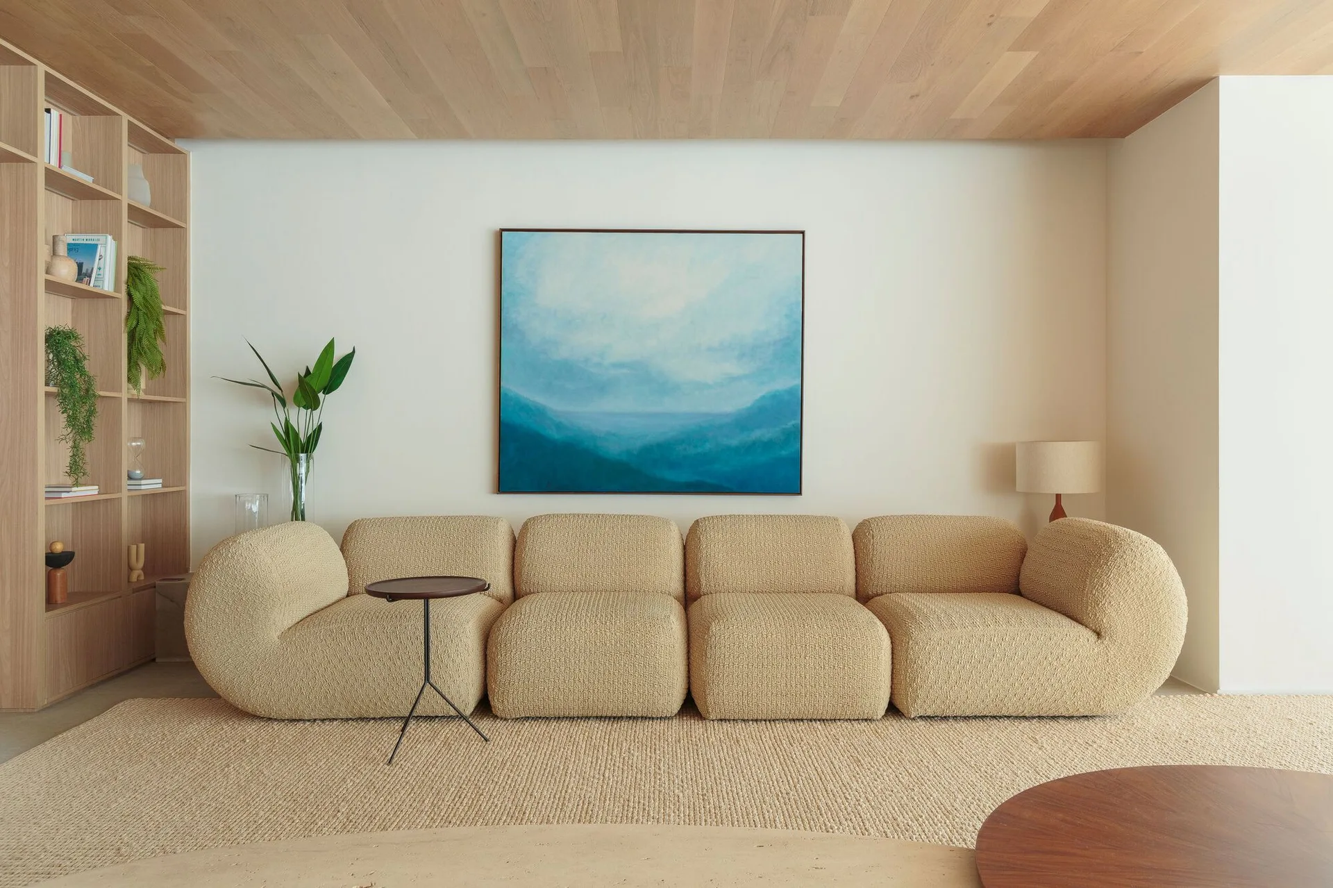 A low-profile modular sofa sits on a woven rug in a living room featuring a wood-planked ceiling and a large blue painting.