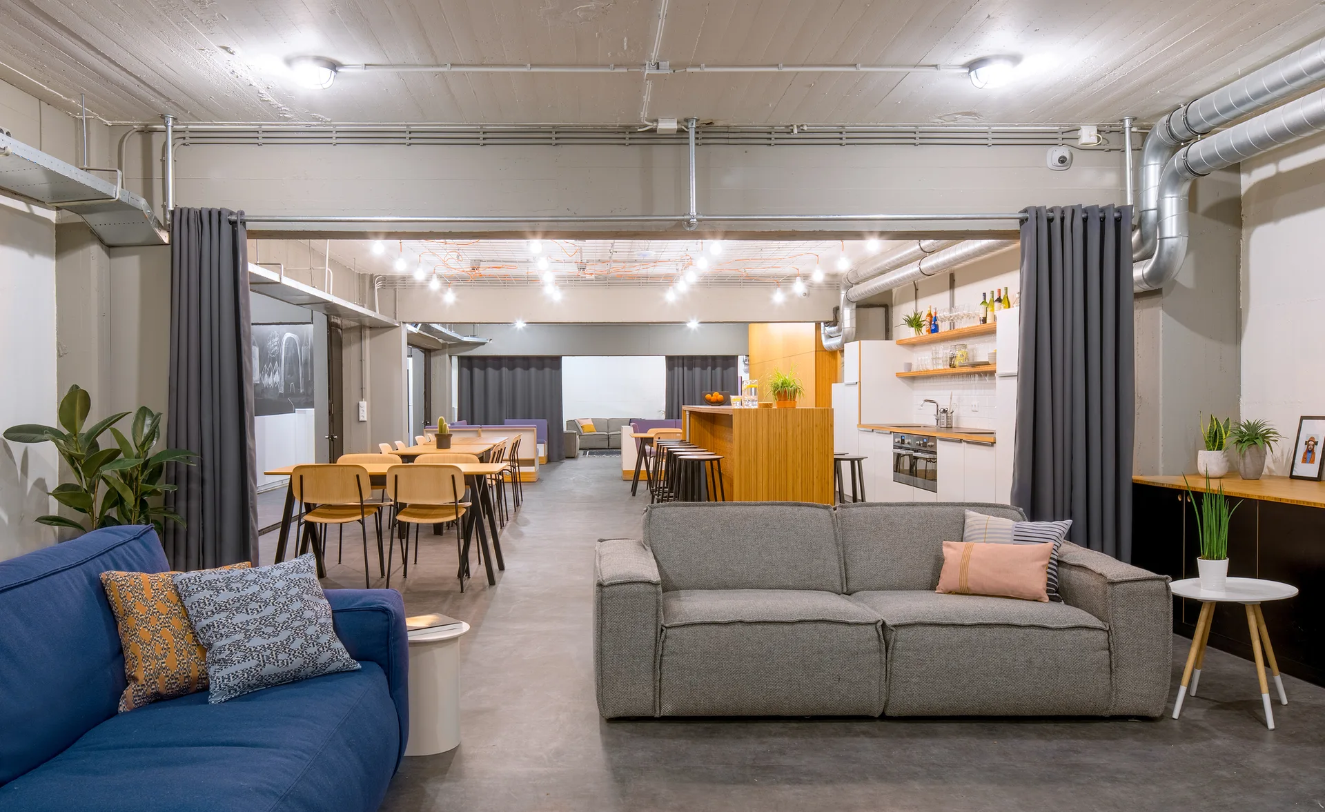 An industrial loft features gray and blue sofas on concrete floors beneath an exposed concrete ceiling with metal pipes and hanging lights.