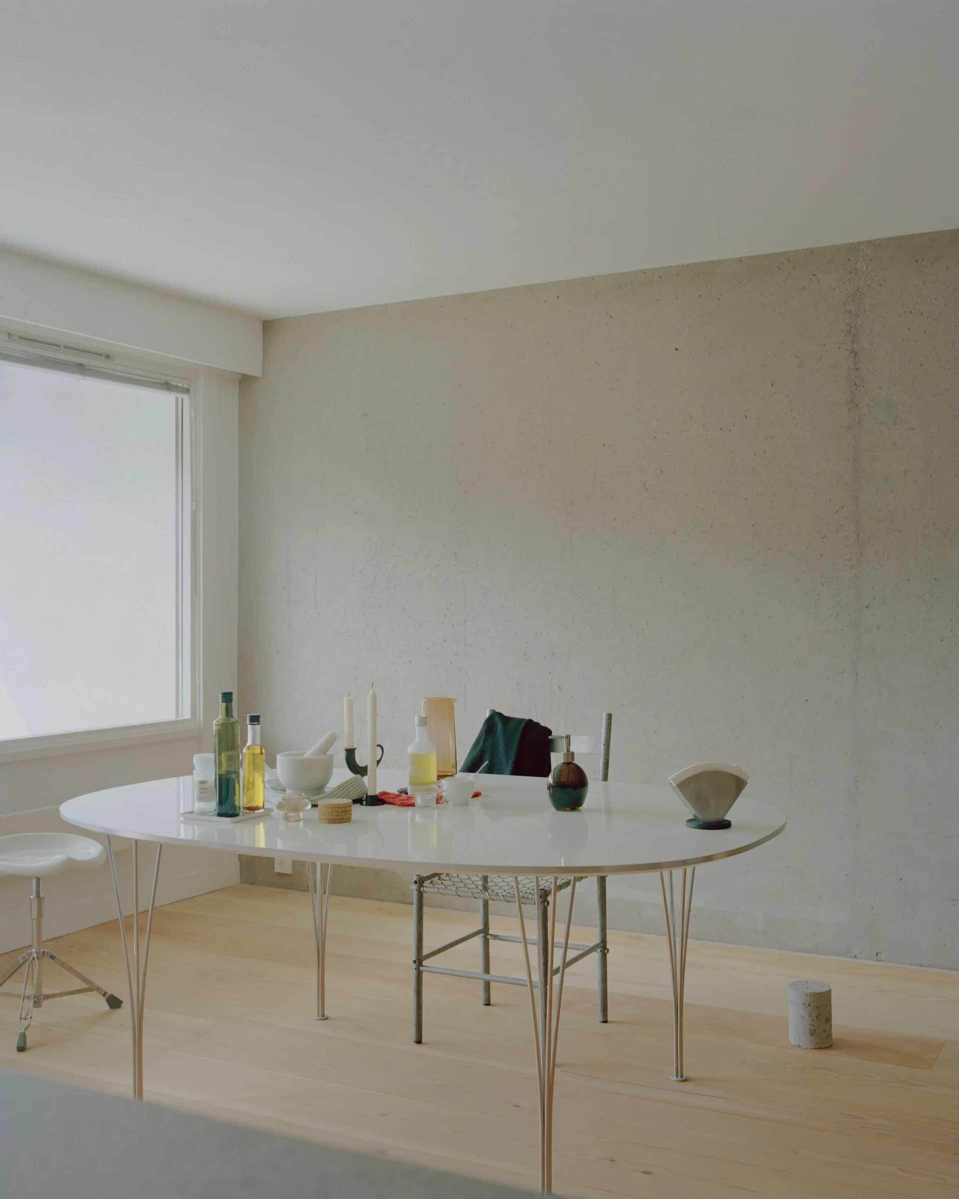 An oval white dining table with slender metal legs sits in a minimalist room with a raw concrete wall and light wood flooring.