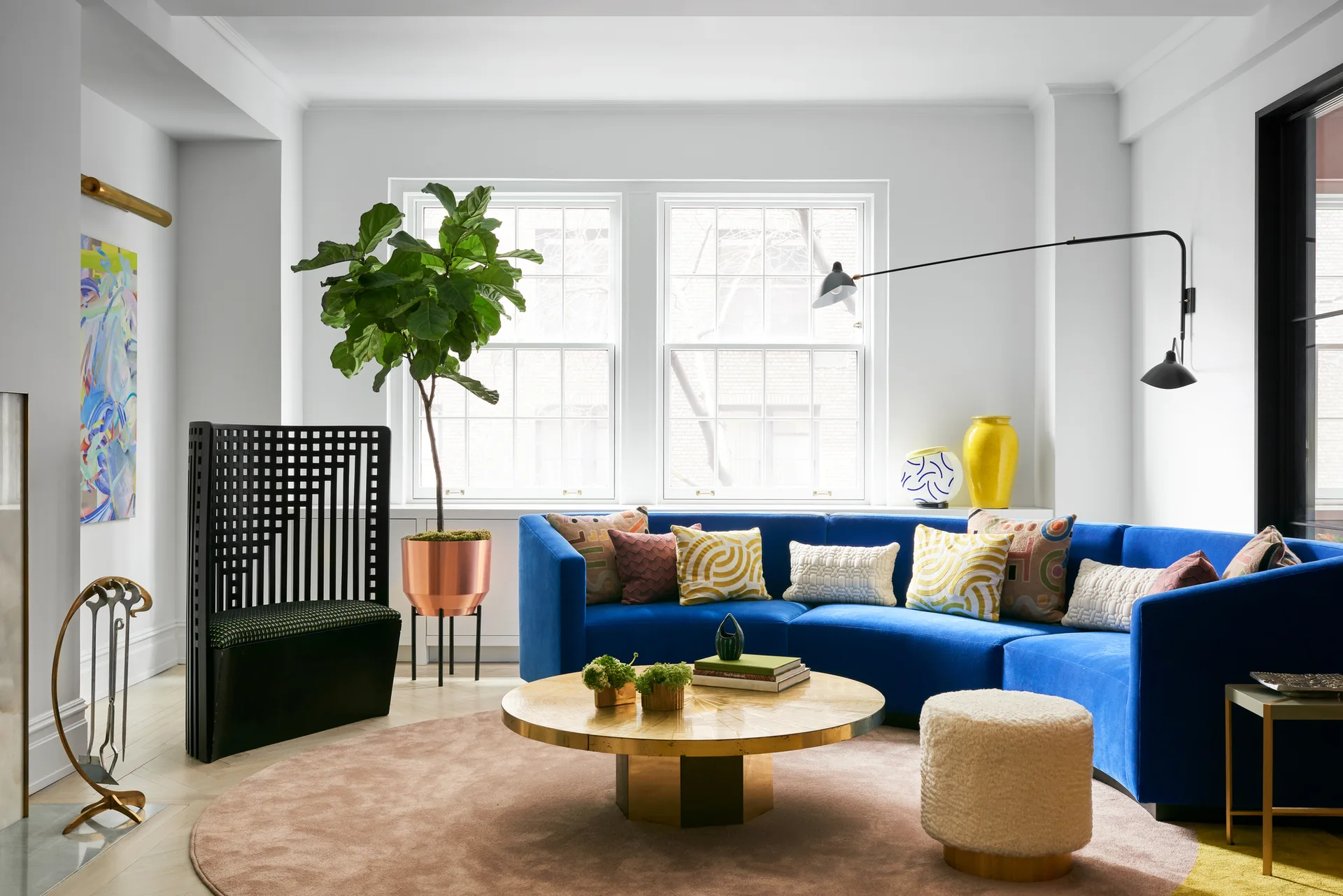 A bright living room features a blue sectional sofa, a round brass coffee table, a tall black slatted chair, and a large pink area rug.