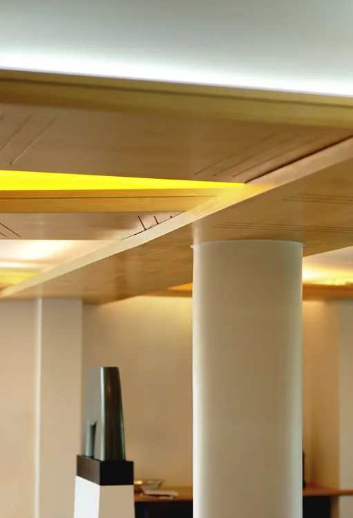 A cylindrical white structural column supports a wood-toned paneled ceiling featuring integrated warm yellow cove lighting.