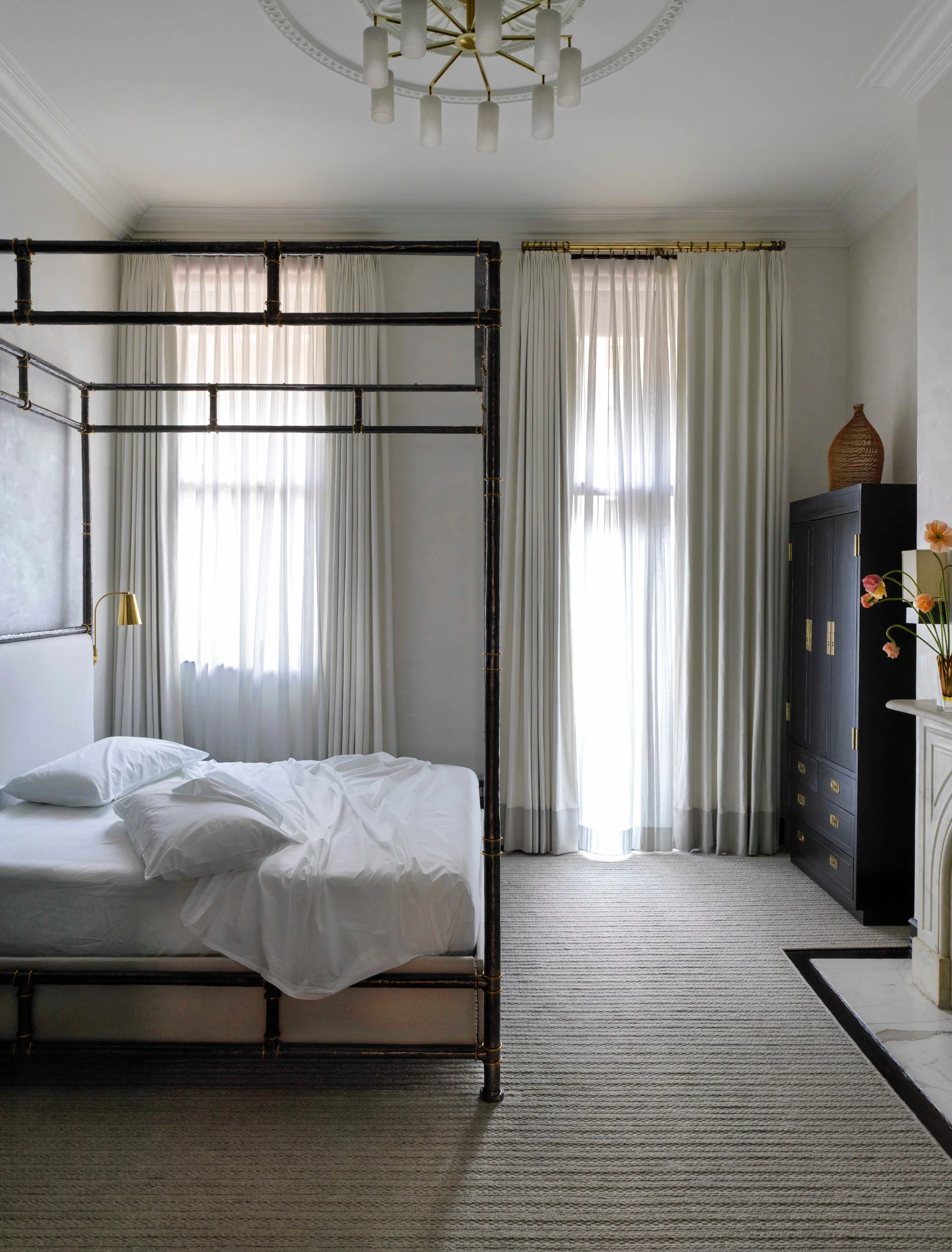 A neutral transitional bedroom features a dark metal canopy bed, floor-to-ceiling cream curtains, a tall black wardrobe, and a ribbed beige carpet.