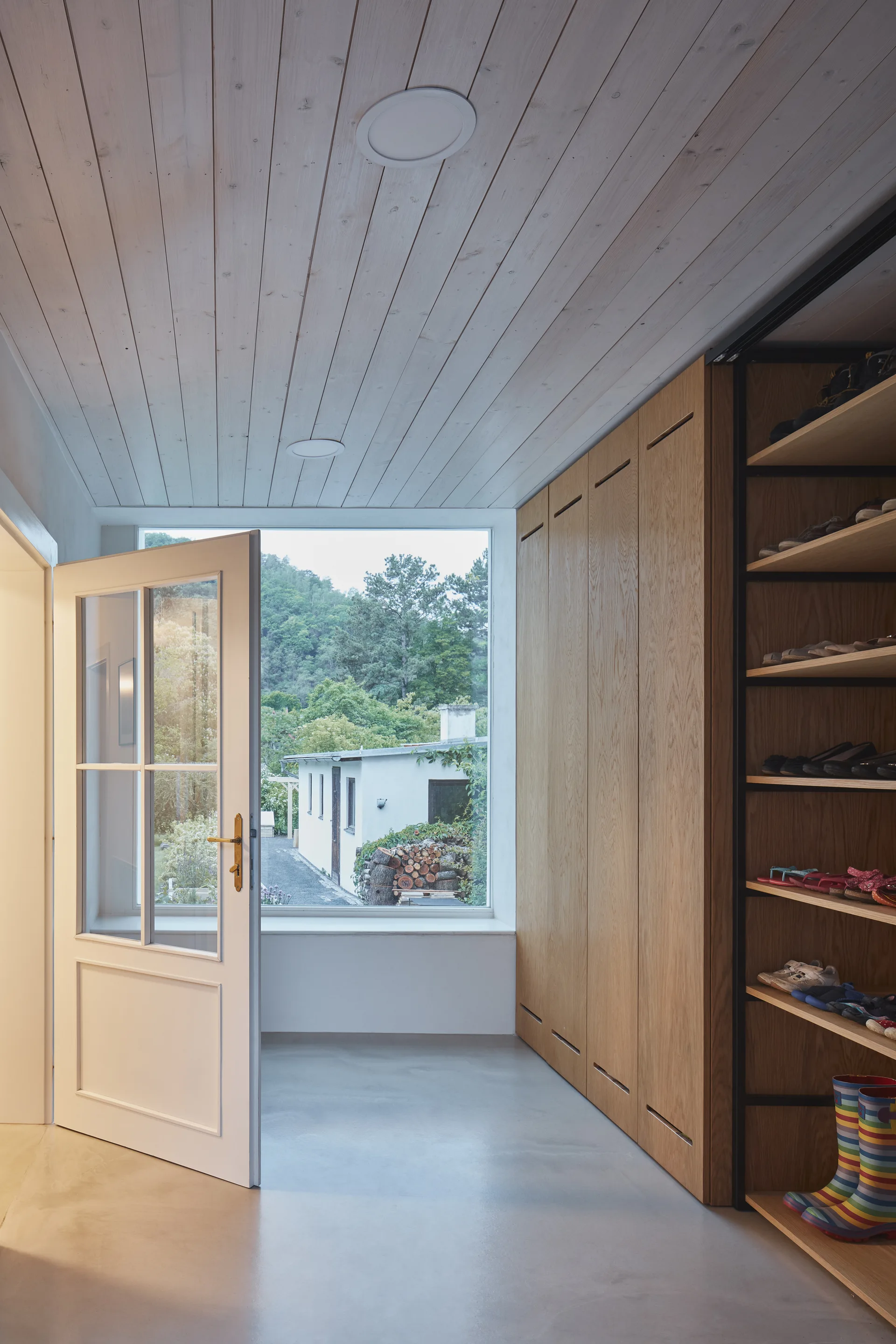 A bright mudroom featuring light oak storage cabinets, open shoe shelves, a white wood plank ceiling, and an open glass-paneled door.
