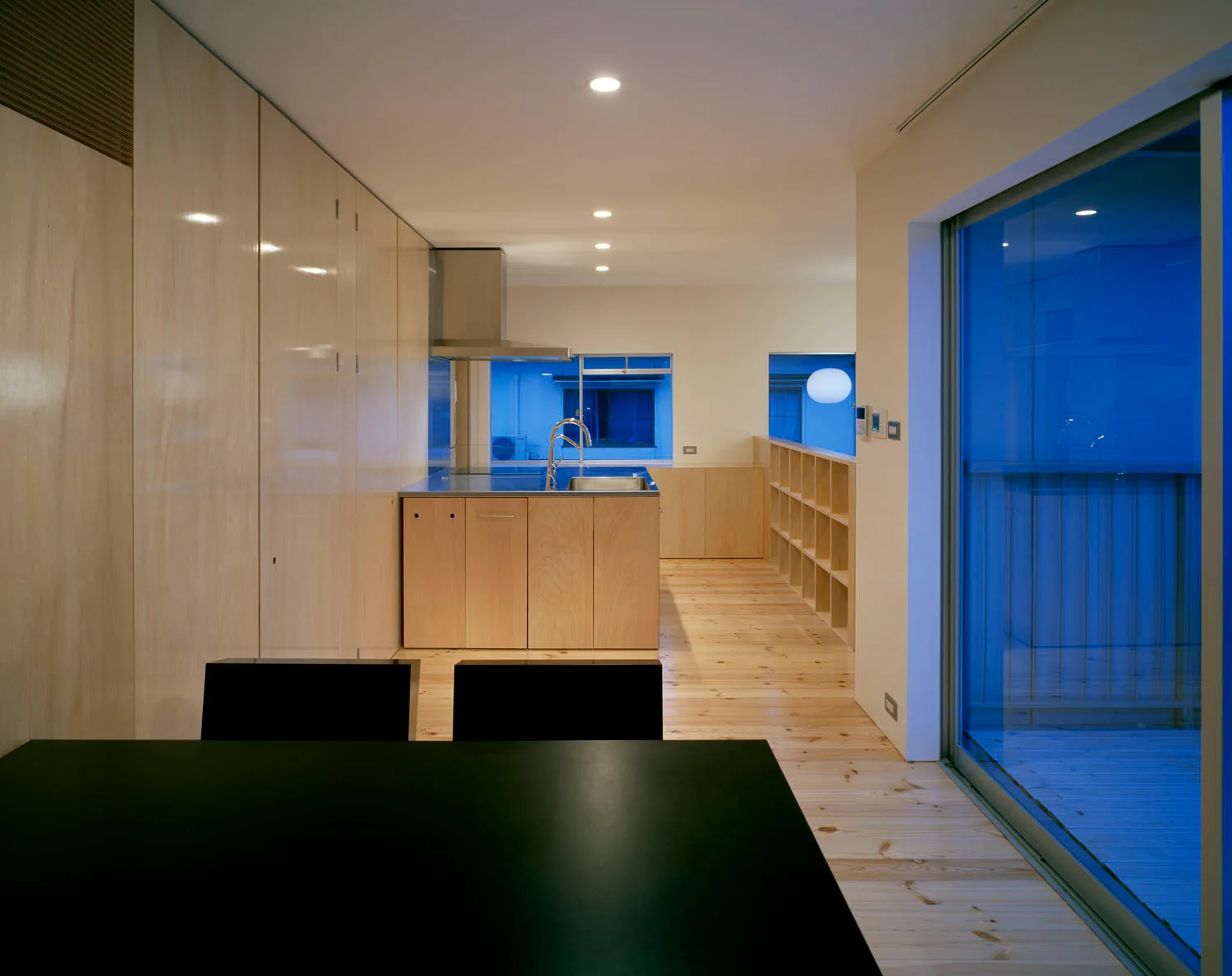 A minimalist kitchen features light wood cabinetry, a matching island, light wood plank floors, and a dark dining table in the foreground.