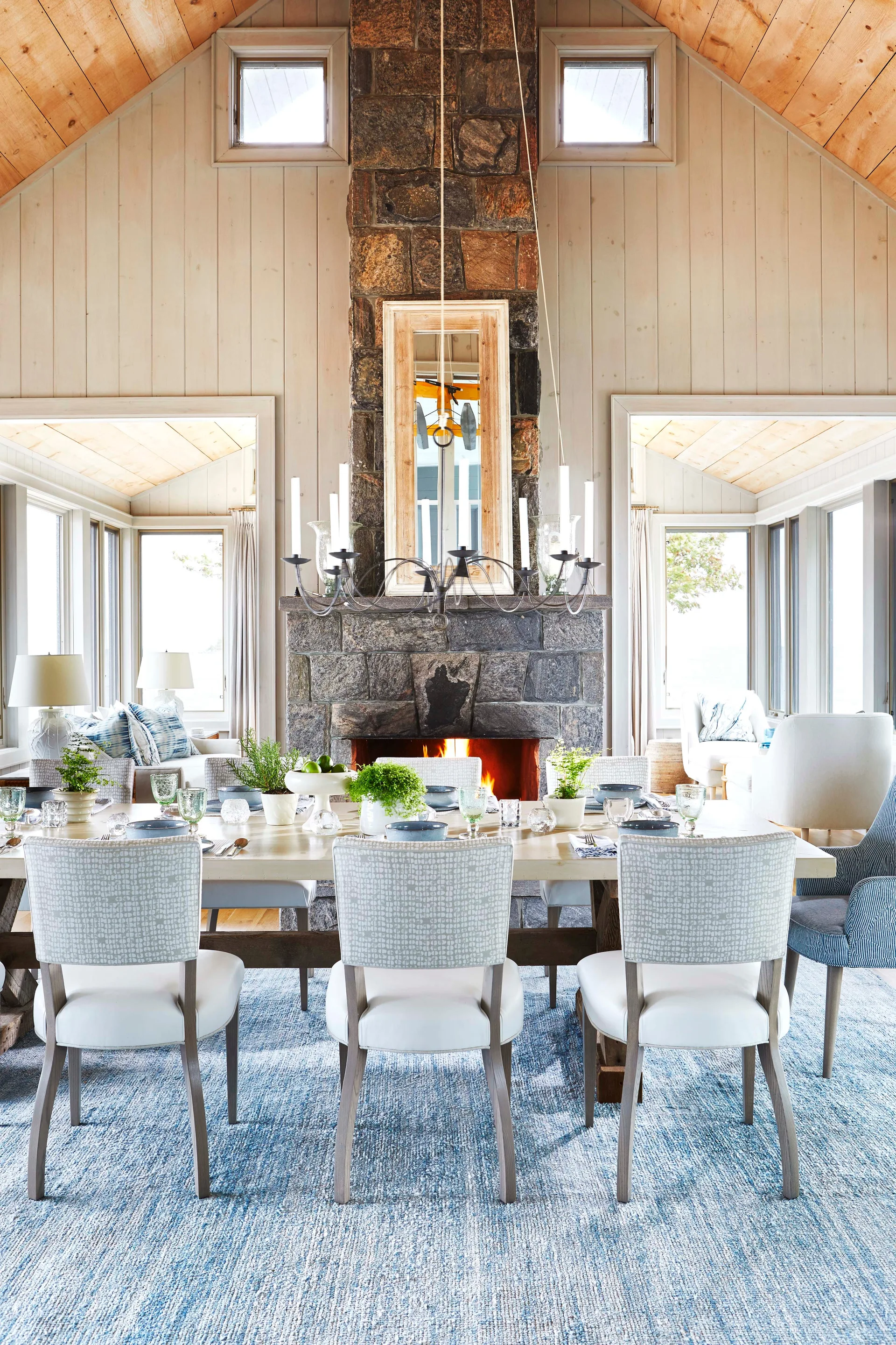 A bright dining room features a long wooden table, patterned chairs, a textured blue rug, and a central stone fireplace.