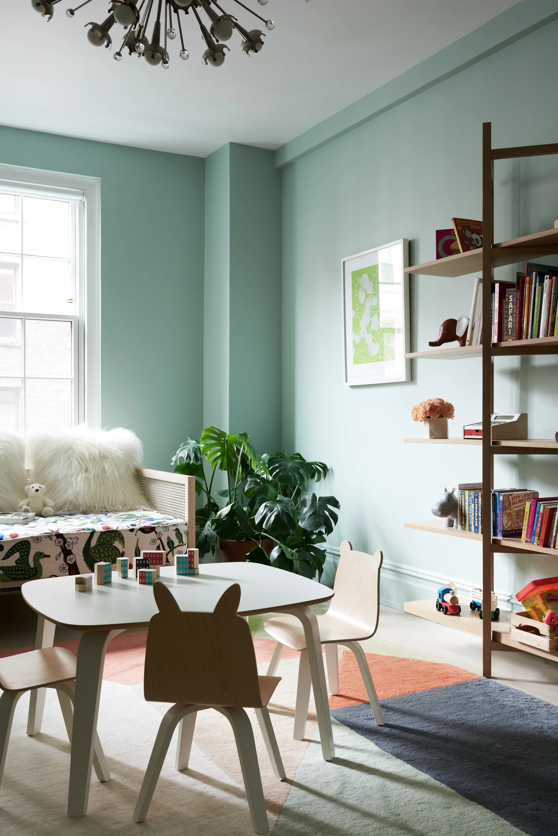 A children's playroom with pastel mint walls, a light wood table with animal-ear chairs, a colorful geometric rug, and a tall open bookshelf.