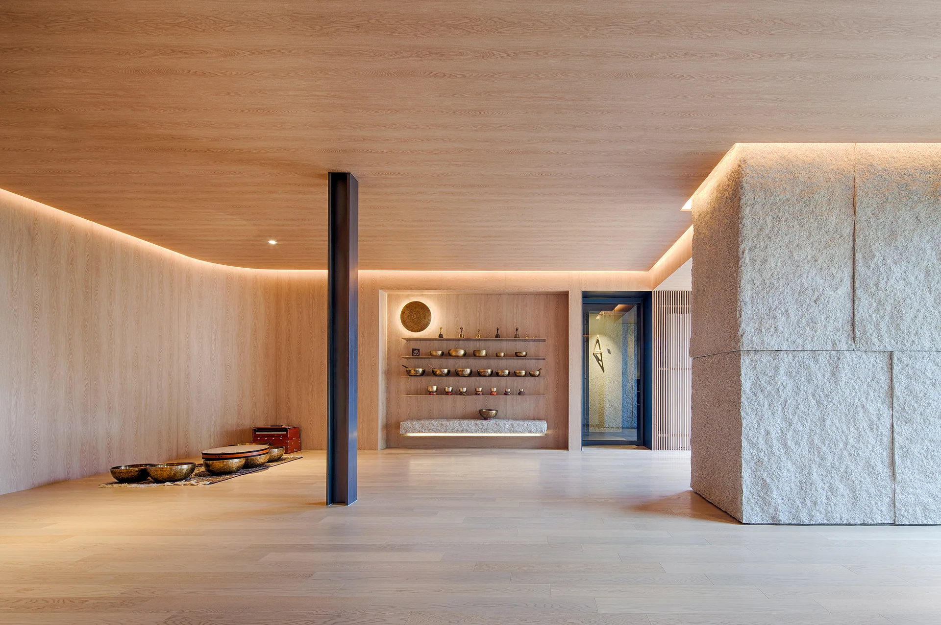 A minimalist wellness room features light wood walls and ceiling, a textured stone pillar, and floating wooden shelves displaying singing bowls.