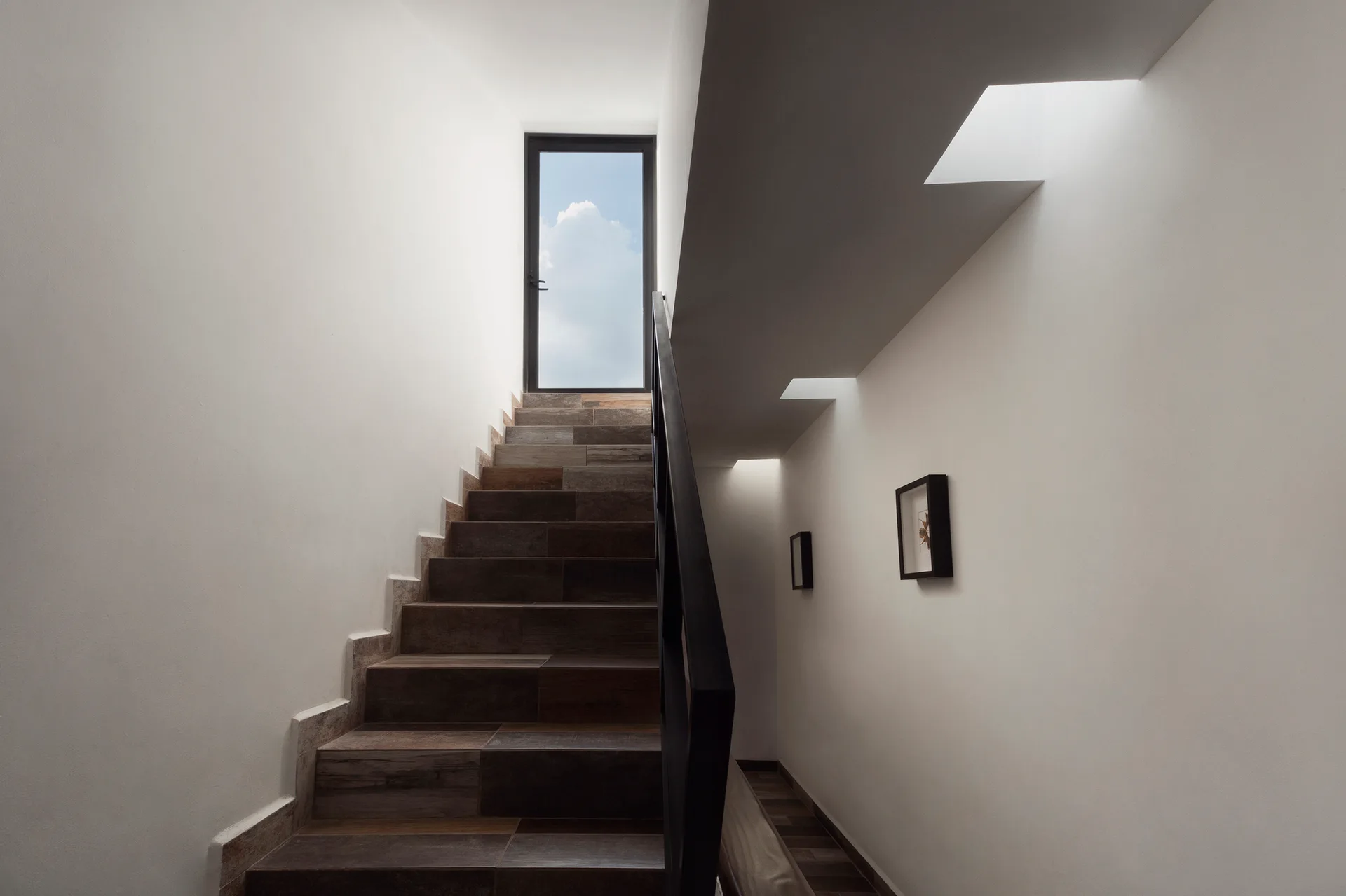 A modern staircase with dark wood-look treads and a black metal handrail rises between white walls toward a glass door framing the blue sky.