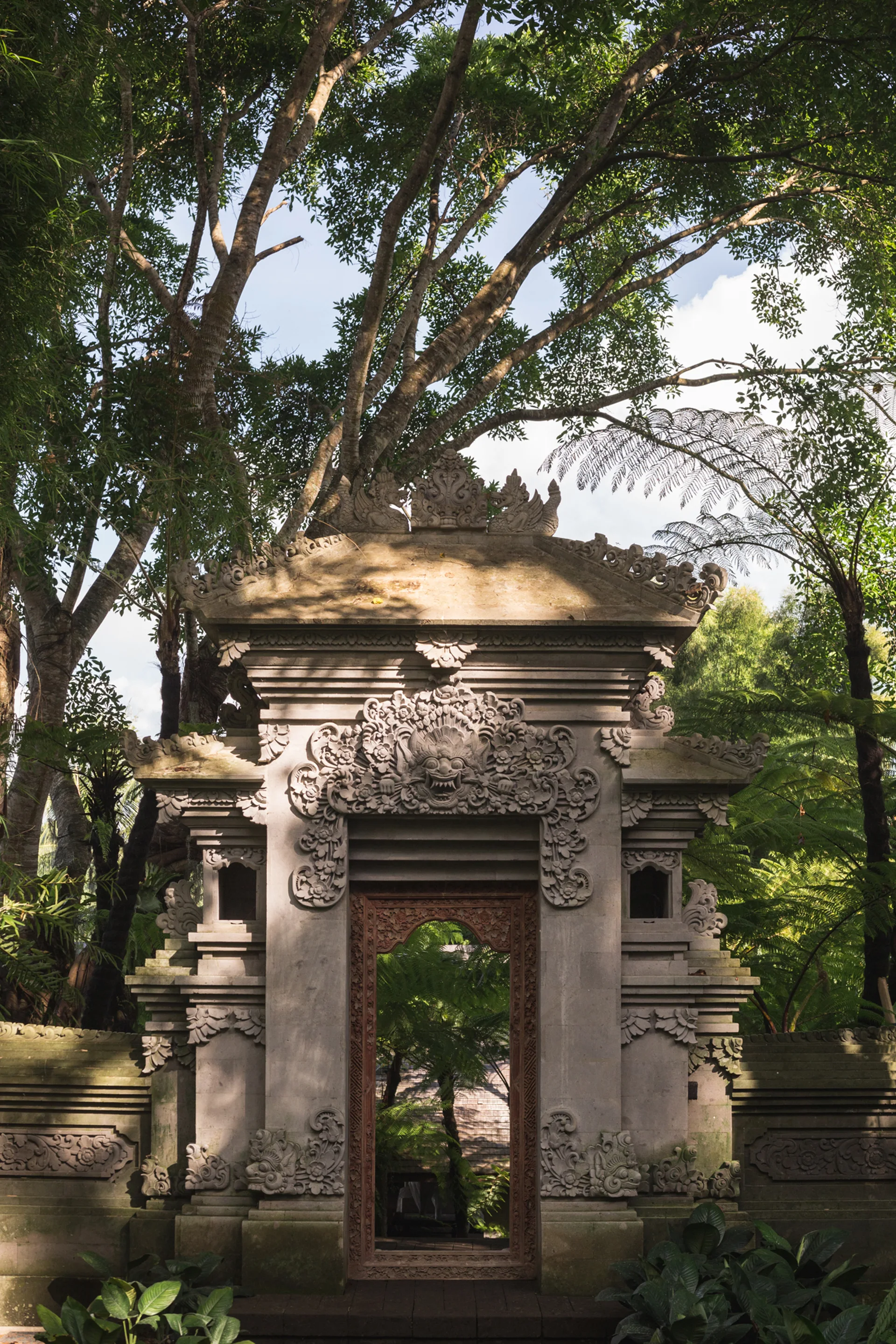 An ornate stone Balinese gateway with intricate carvings is surrounded by tall trees and lush green plants in a garden.