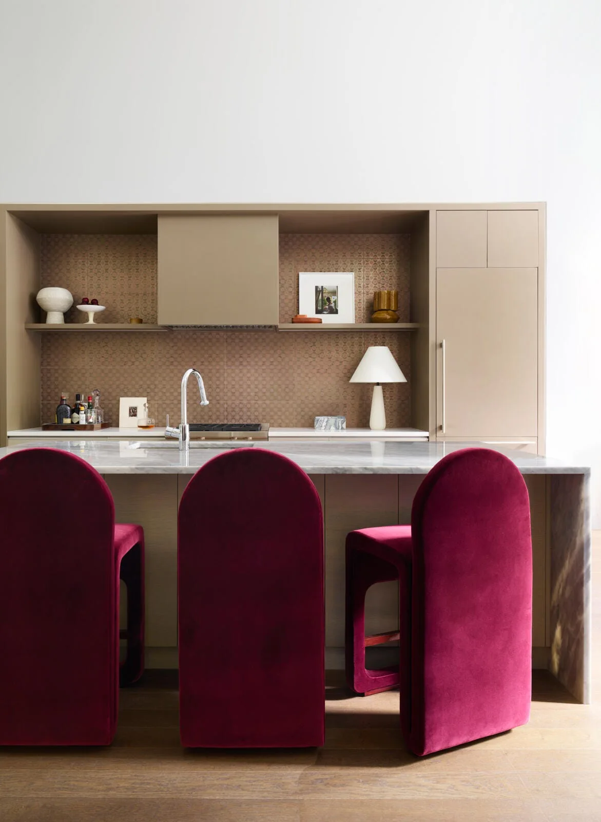 Three magenta velvet bar stools sit at a marble waterfall island in a modern kitchen with beige cabinets.