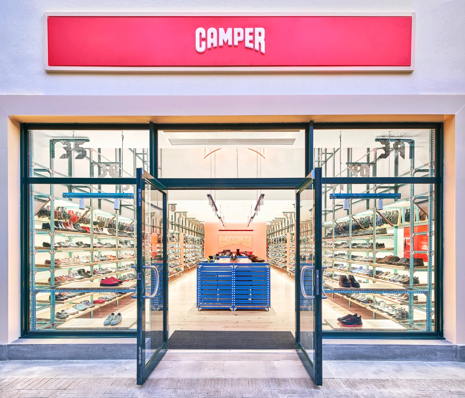The glass entryway of a shoe store reveals rows of industrial metal shelving holding shoes, a central blue unit, and a red brand sign above.