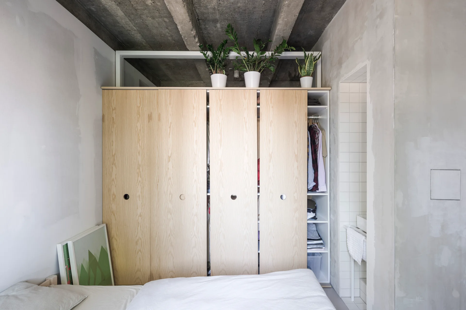 A bedroom features a light wood wardrobe with circular cutouts standing beneath an unfinished concrete ceiling.