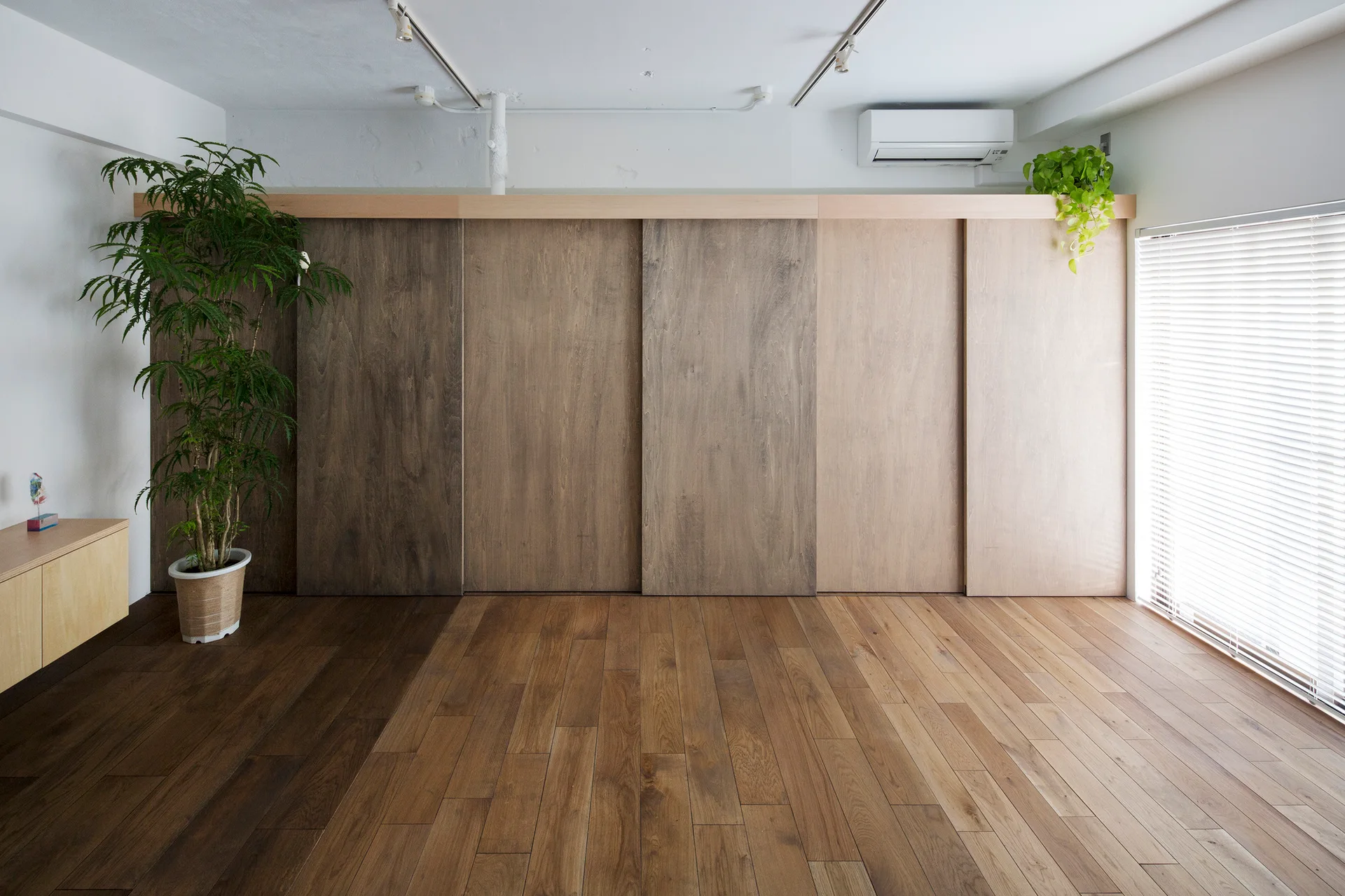 A minimalist room featuring large wooden sliding wall panels, medium-toned hardwood flooring, and a large window with white blinds.